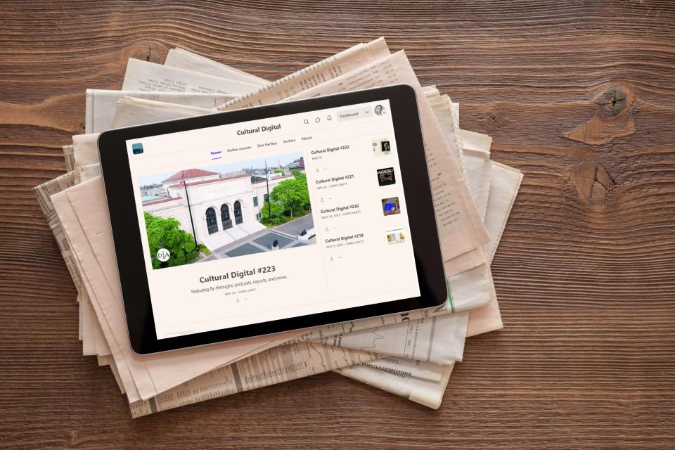 Tablet showing the Cultural Digital newsletter, issue #223, resting on a stack of newspapers on a wooden table.