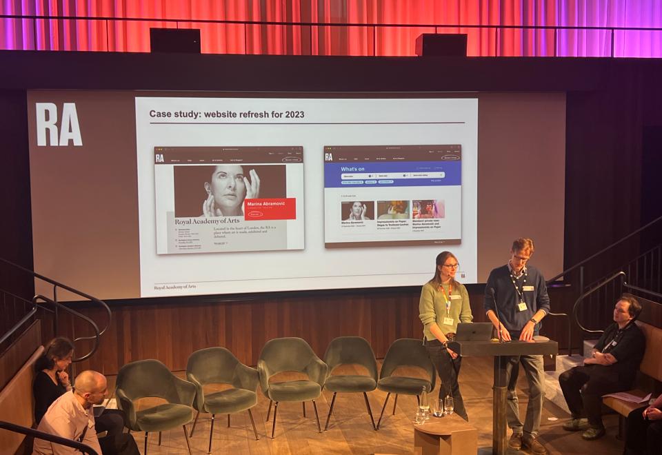 Two speakers at a lectern beside a screen showing a Royal Academy of Arts slide, 'Case study: website refresh for 2023', at the MCG Conference.