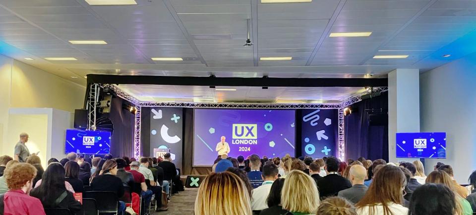 Delegates seated facing a speaker on stage at UX London 2024, with the UX London logo on the screens.