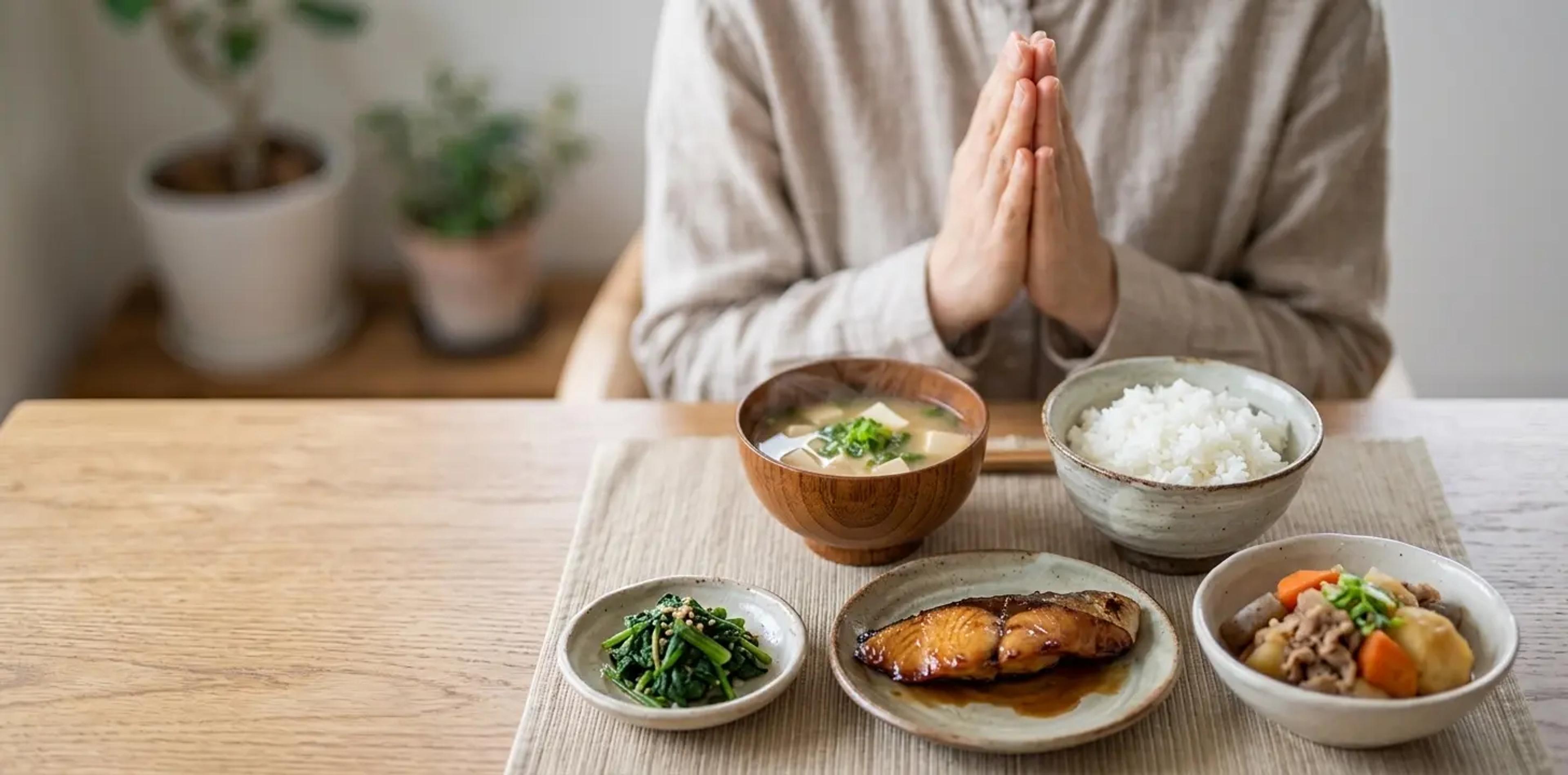 A person clasping hands in gratitude before a traditional Japanese meal of rice, miso soup, and side dishes, illustrating the concept of Itadakimasu.