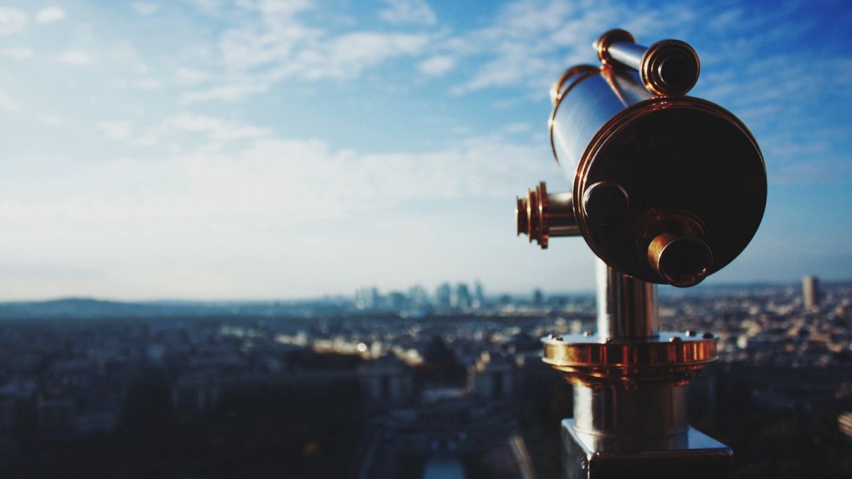 A telescope on top of a high viewpoint is in focus, overlooking the blurry skyline in the distance under a bright blue sky with patchy clouds. A telescope on top of a high viewpoint is in focus, overlooking the blurry skyline in the distance under a bright blue sky with patchy clouds.