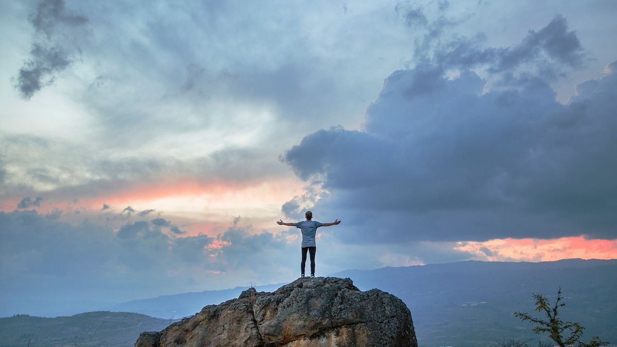 Joshua Earle's photo of the back of a man with arms out wide on top of a boulder that is overlooking a cloudy landscape with the sun setting. Joshua Earle's photo of the back of a man with arms out wide on top of a boulder that is overlooking a cloudy landscape with the sun setting.