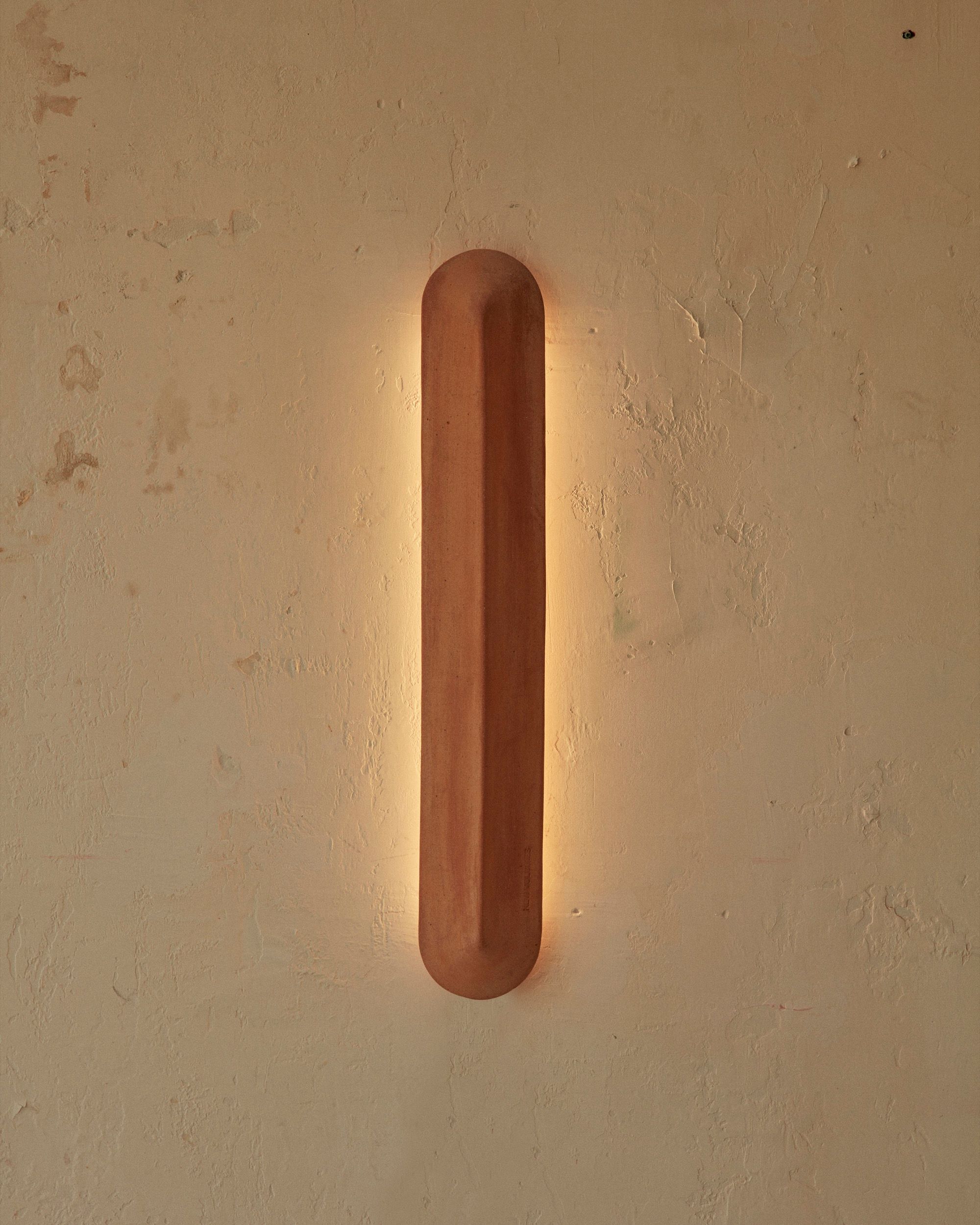A Pinch 900 elongated wall light made of terracotta, against a white brick background. Light is illuminating the wall it is fixed to