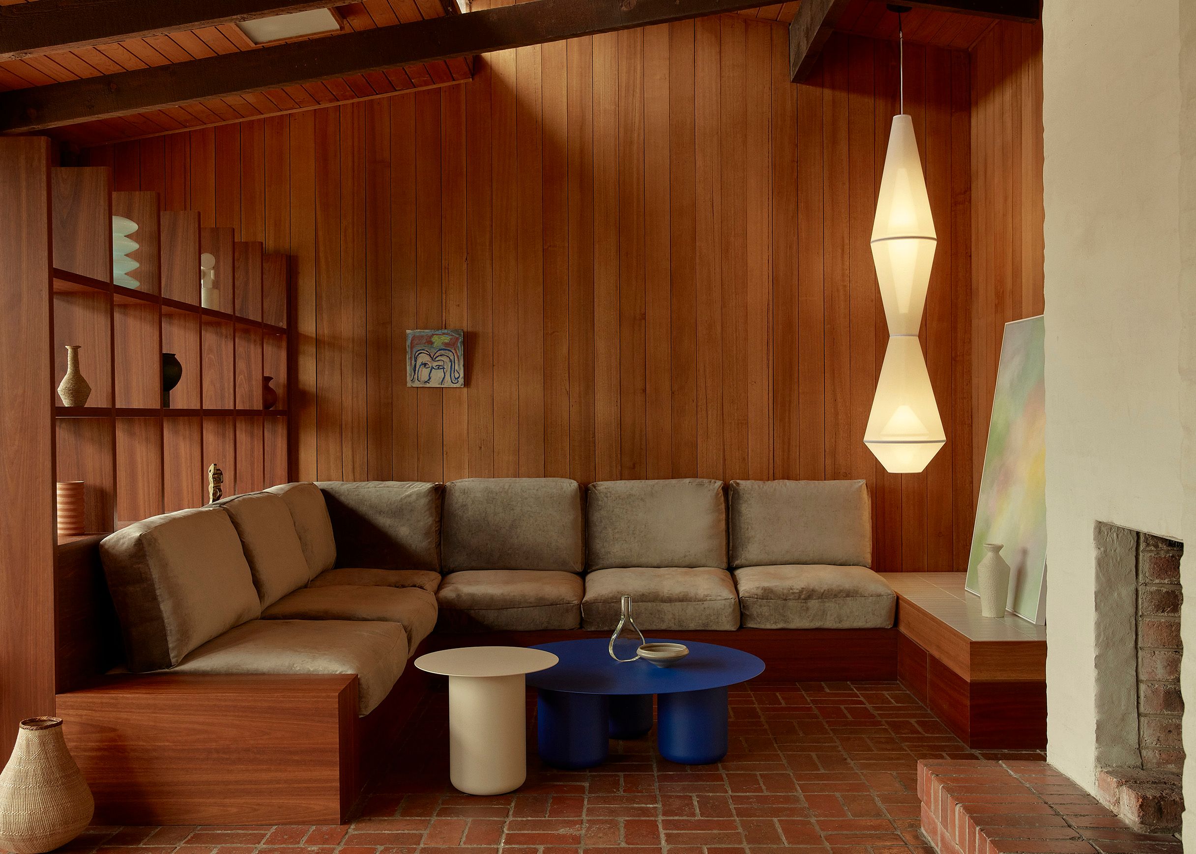 A large mesh fabric illuminated pendant light hangs in a living room with timber floors and walls. The Sofa is soft brown and there is colourful artwork and coffee table