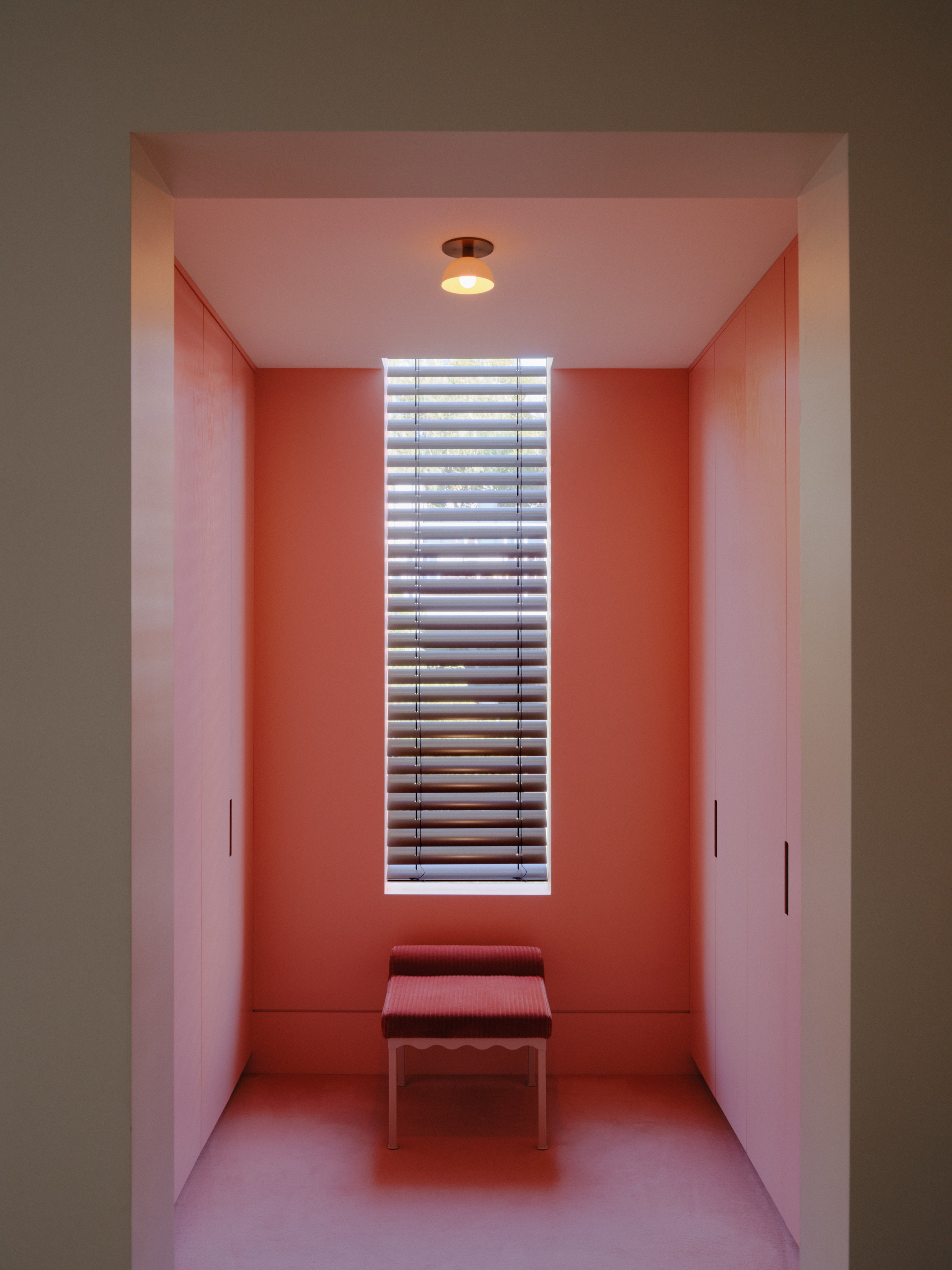 A walk in wardrobe with pink walls and a tall window in the centre of the wall. Under this window is a Bellini upholstered bench