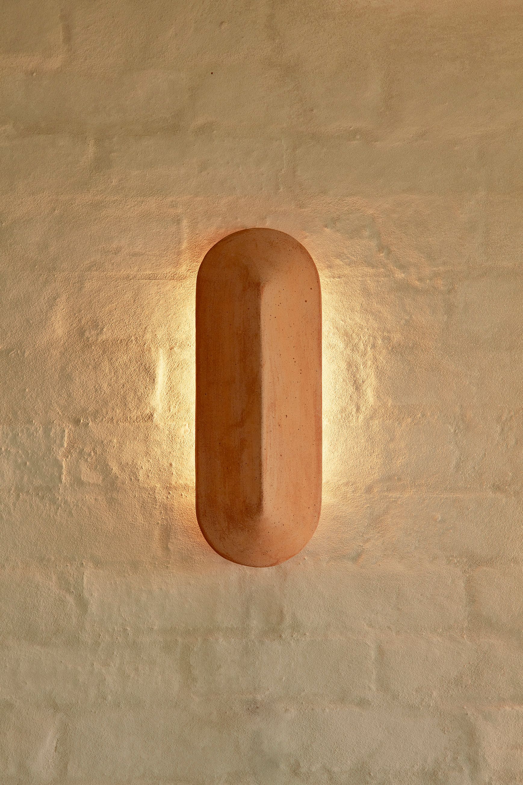 A Pinch 400 elongated wall light made of terracotta, against a white brick background. Light is illuminating the wall it is fixed to