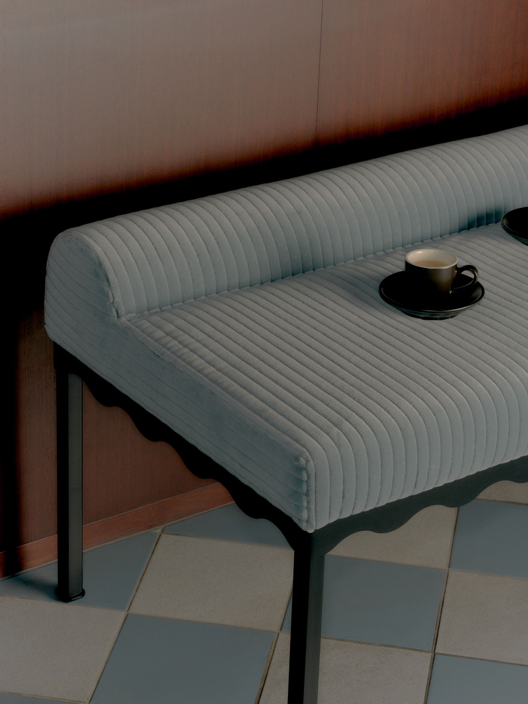 A pale blue wide corduroy upholstered bench in a dimly lit room with blue and white tile flooring