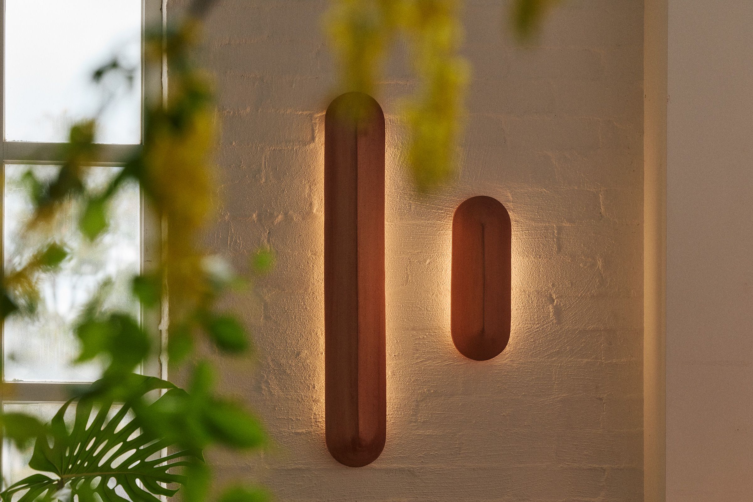 Two terracotta Pinch Wall Lights glowing on a white rendered brick wall with greenery in the foreground
