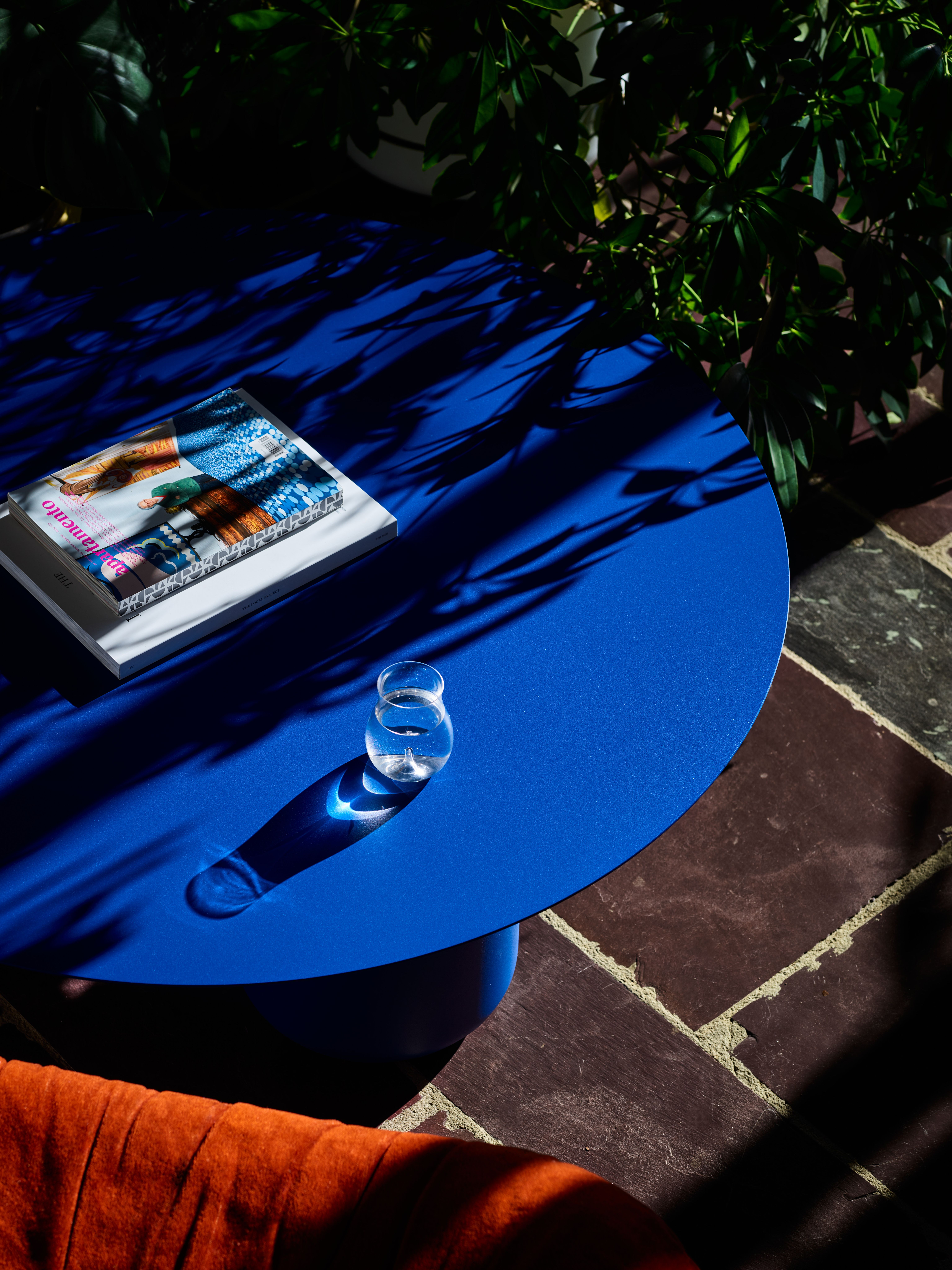 A vivid blue Sequence coffee table with sunlight creating shadows from a plant. There are magazines and a glass of water on the table and a red velvet chair next to it. 