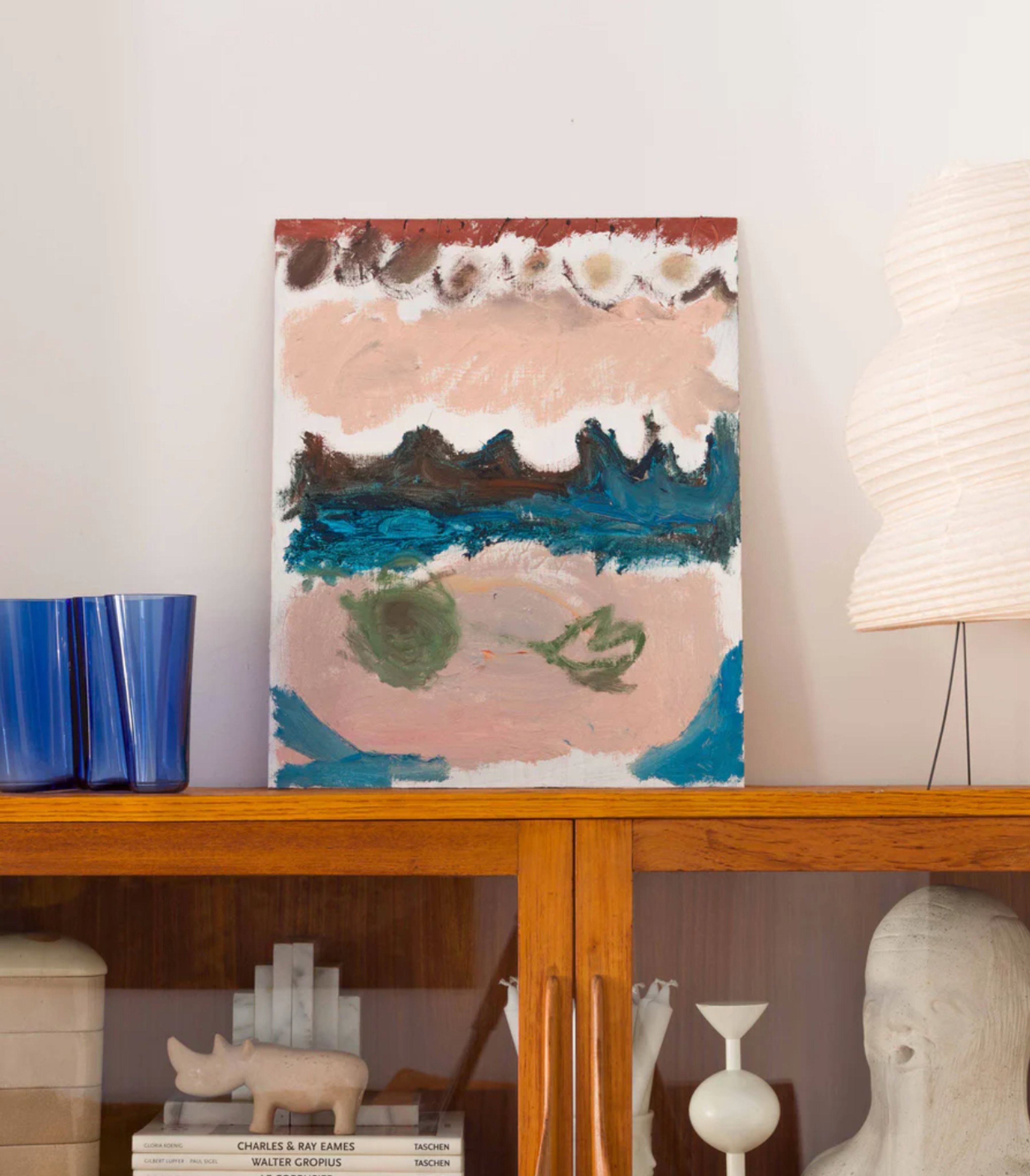 An abstract painting sitting on the top of a wooden cabinet with a lamp and a blue vase