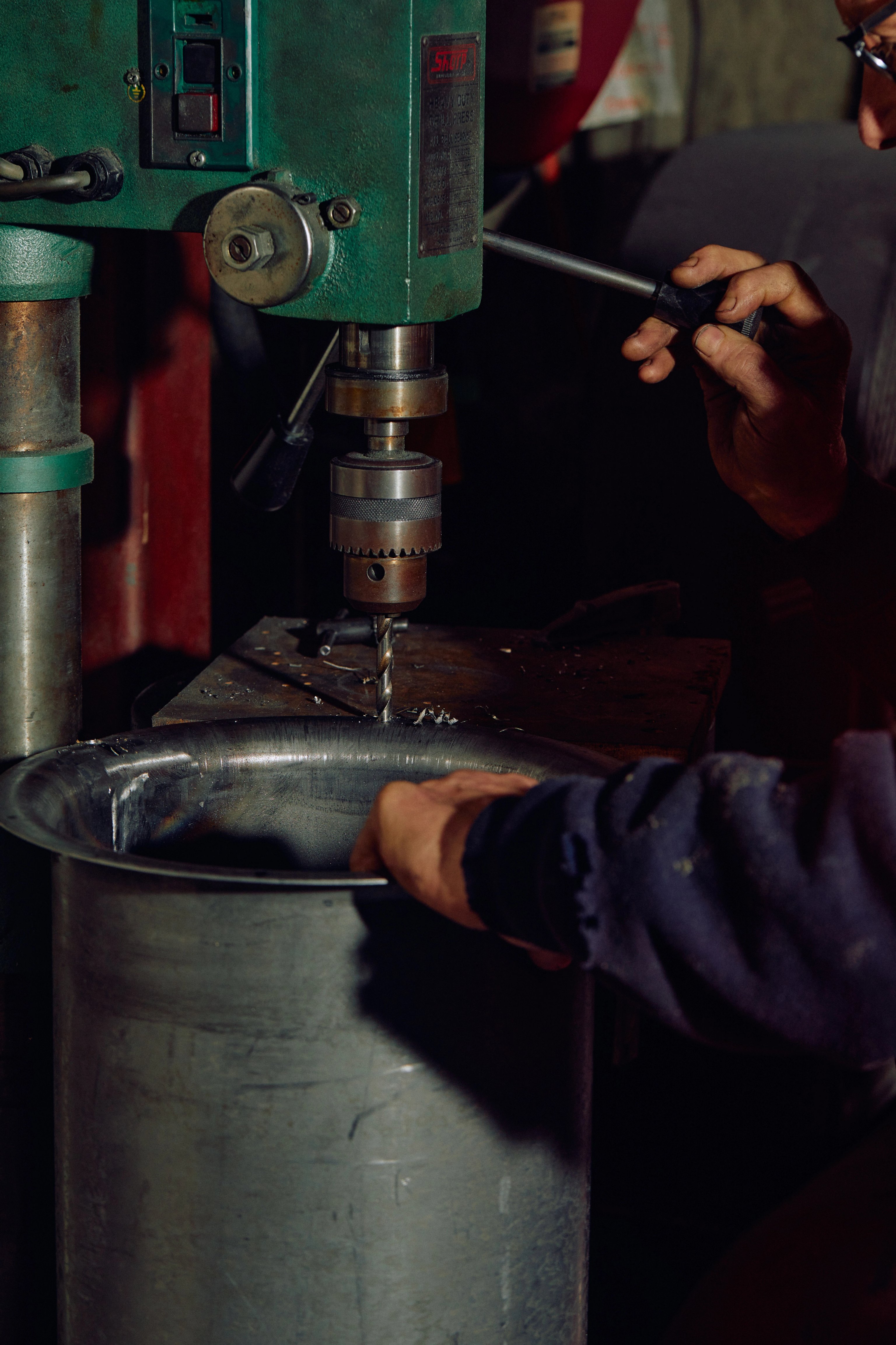 A pair of hands drilling holes into a metal drum with an old industrial machine