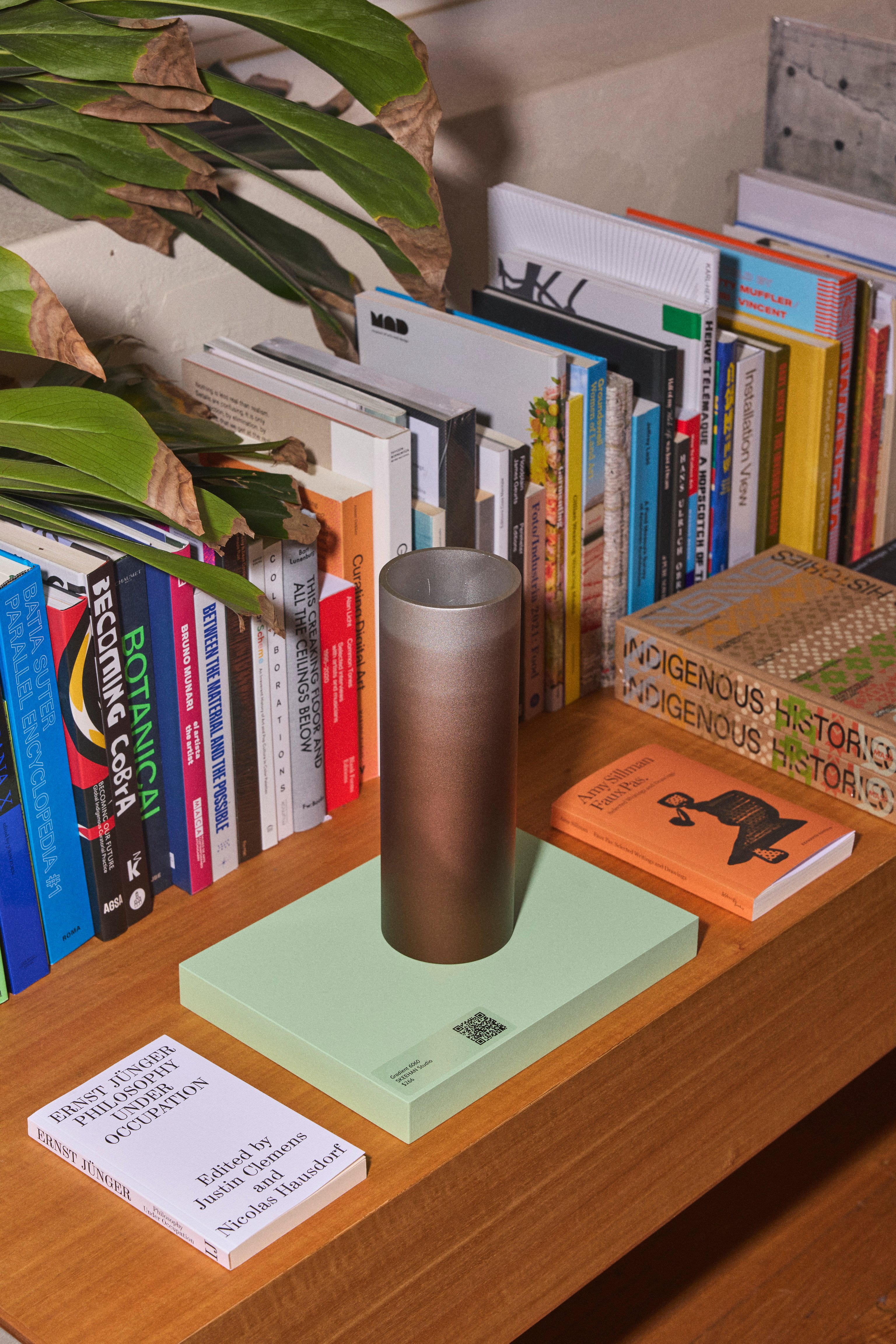 A gradient bronze and silver metal vessel sitting on a pale green book on a bookshelf