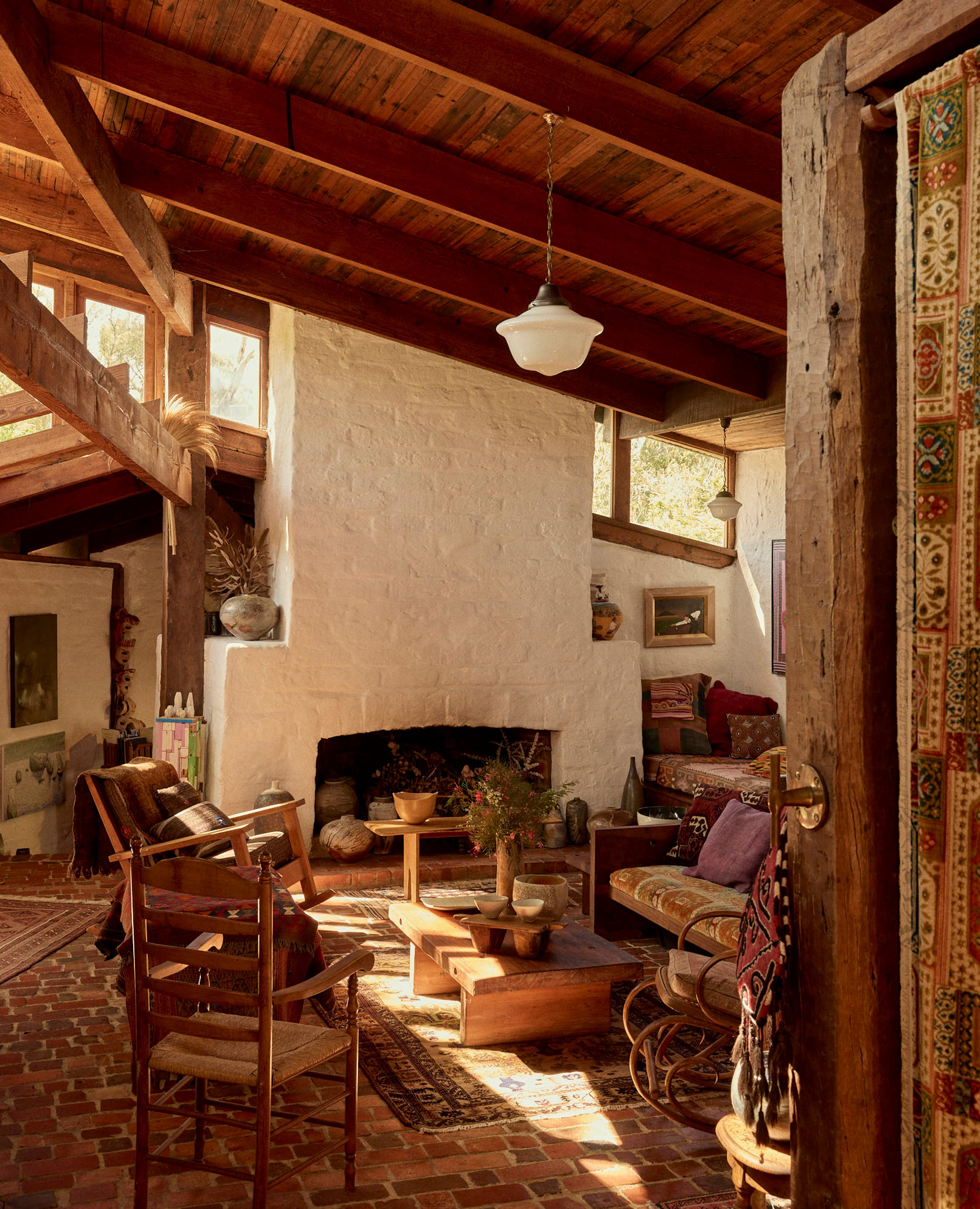 An eclectic home living room with a large open fire, brick floors and timber ceilings. The room is layered with textured furniture and objects throughout and the warm sunlight spills through the high windows.