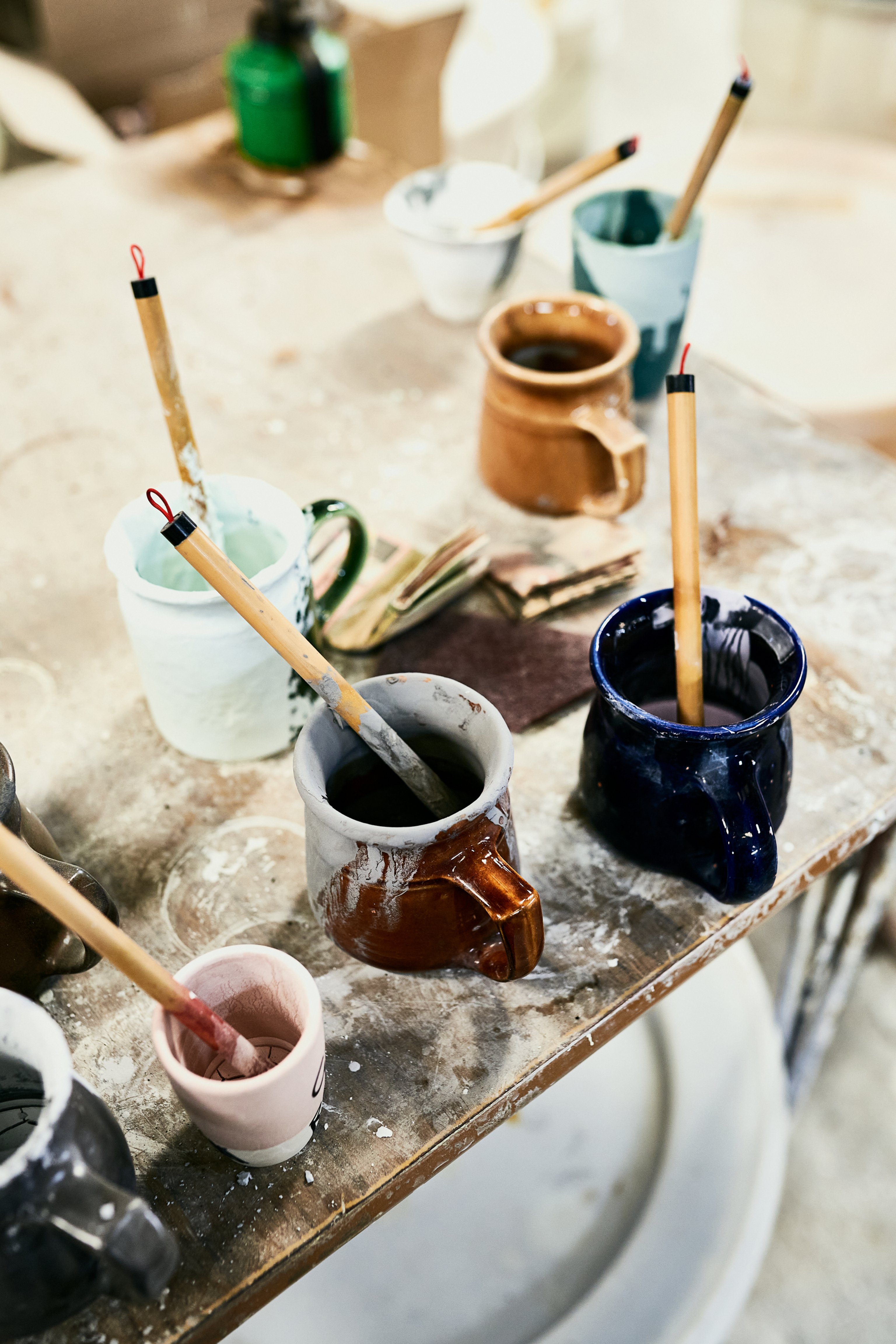A series of coloured mugs sit on a timber workbench with brushes and glaze in them.