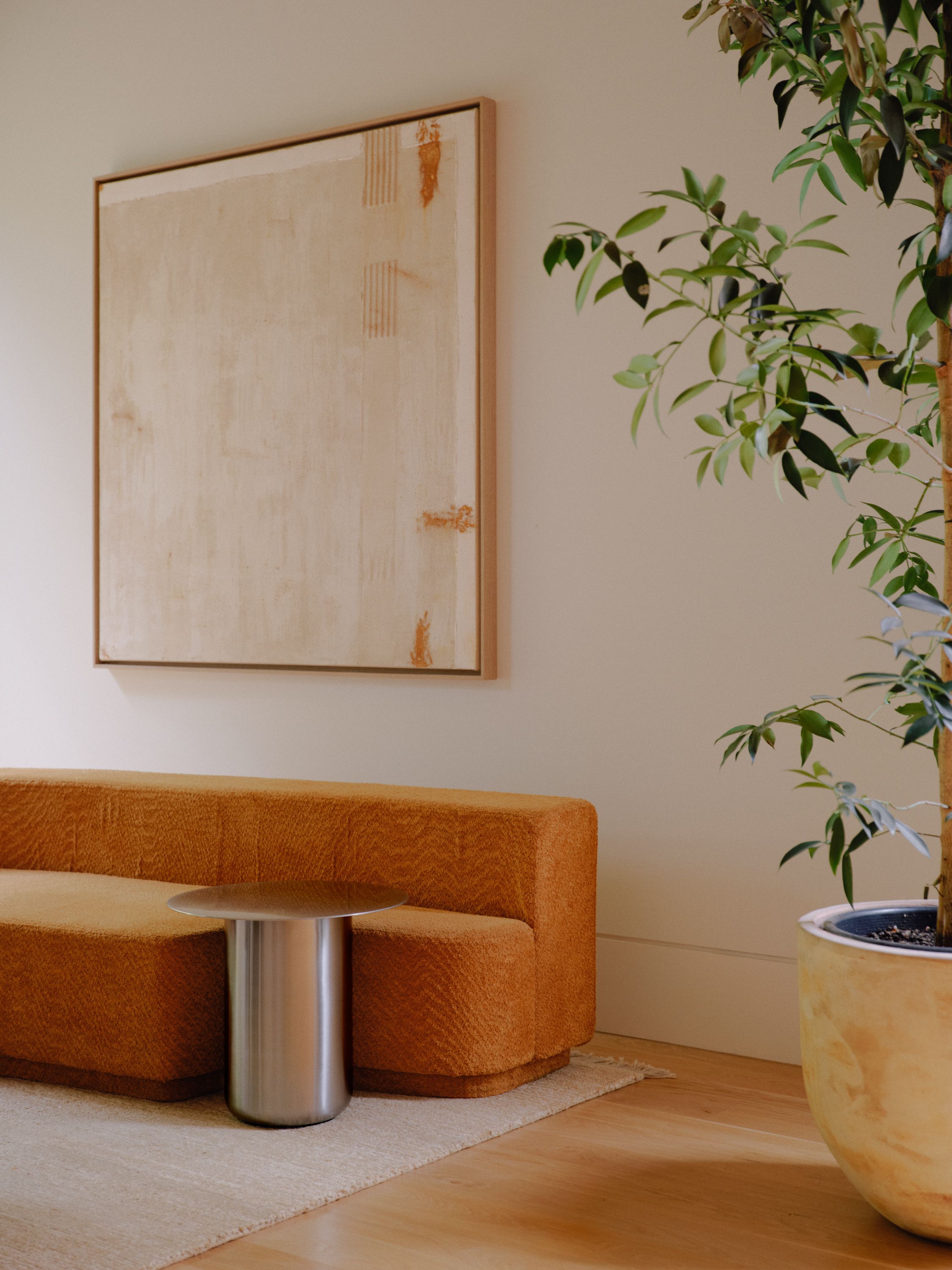 A polished satin stainless steel side table is sitting in a living room with an orange sofa and neutral coloured artwork.
