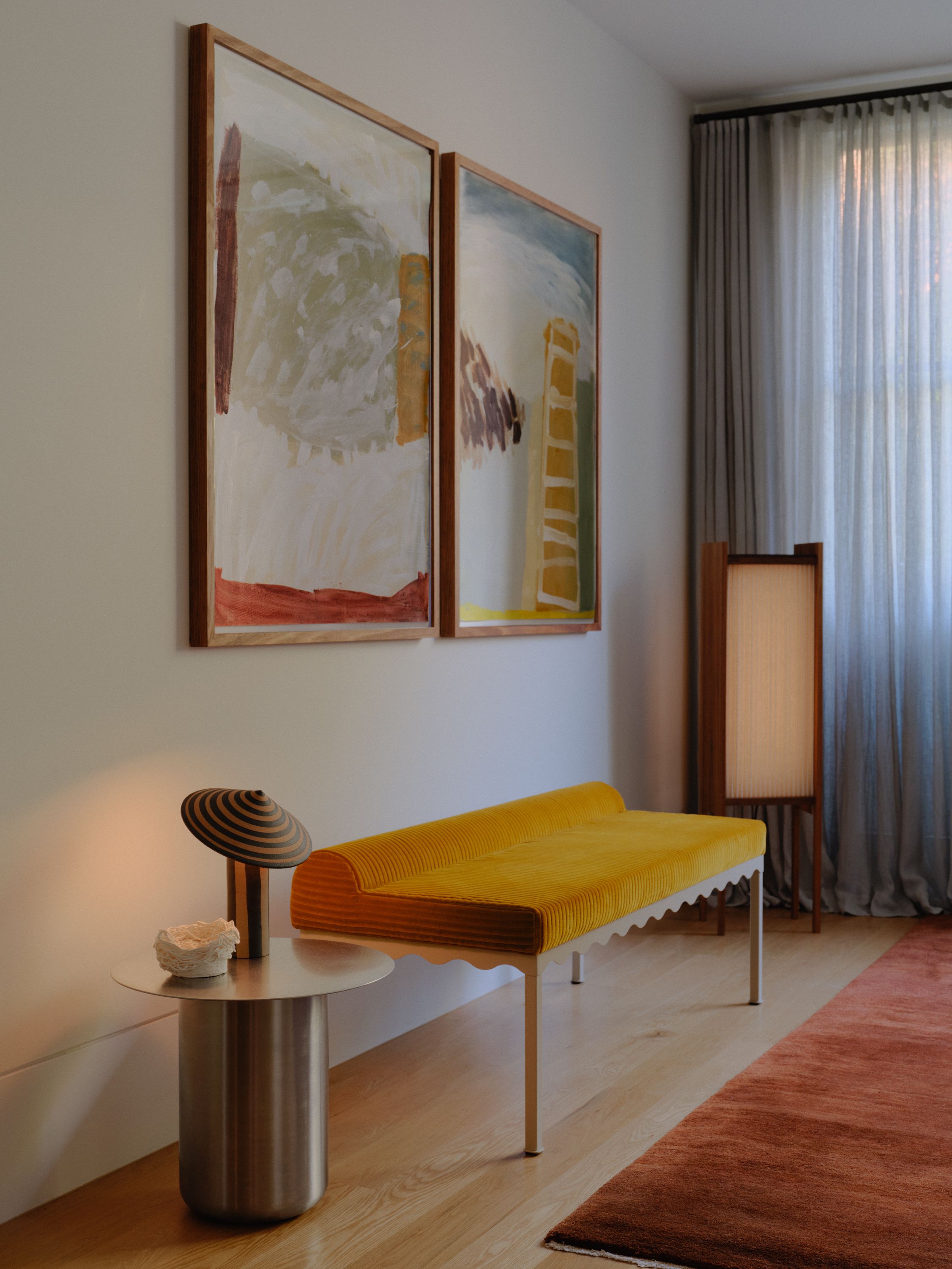 A yellow corduroy upholstered Bellini bench seat in a room with curtains and artwork. The bench sits next to a stainless steel side table and some crafted lighting