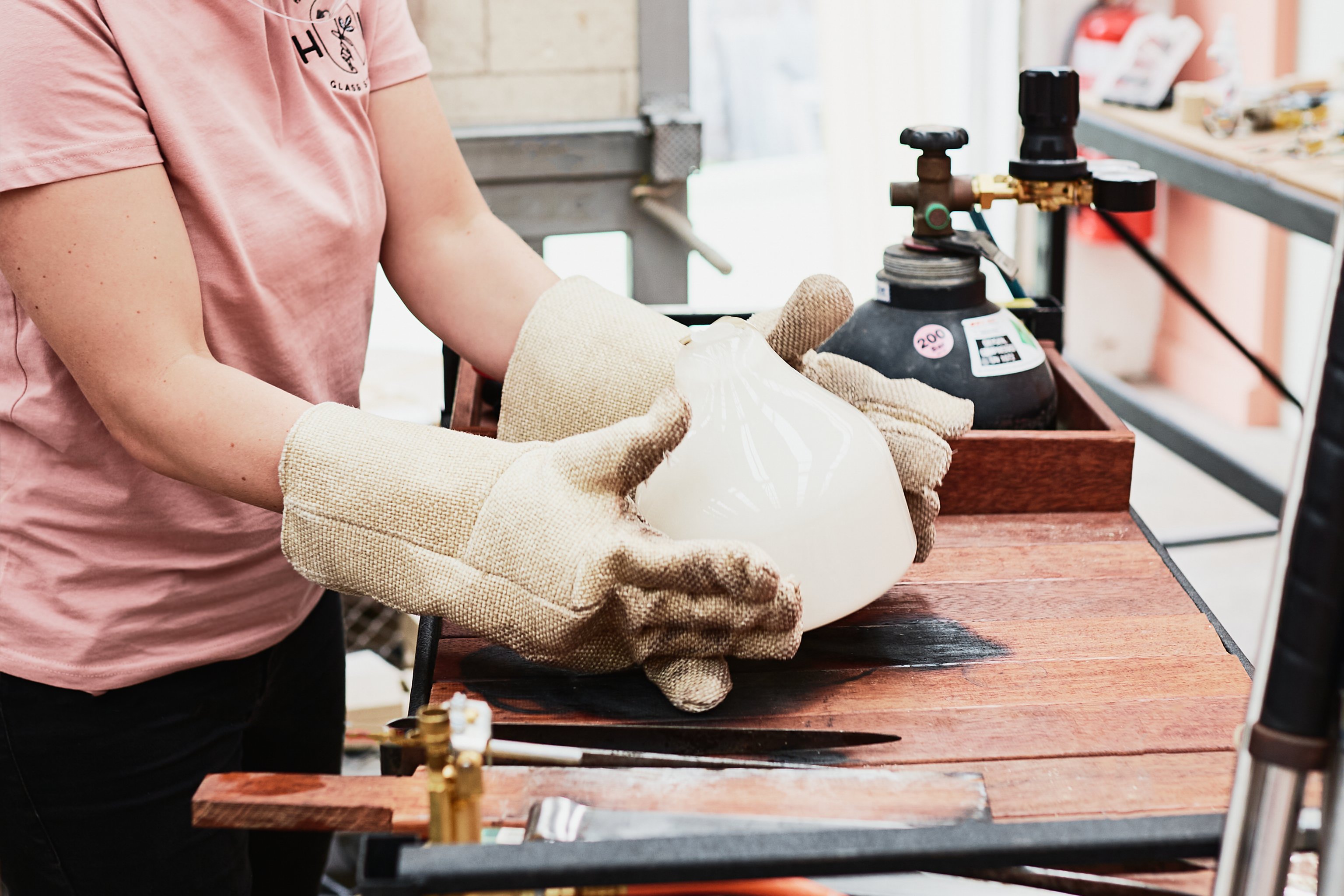 A woman wearing large white gloves holds a piece of white glass above a timber bench with a gas bottle behind it.