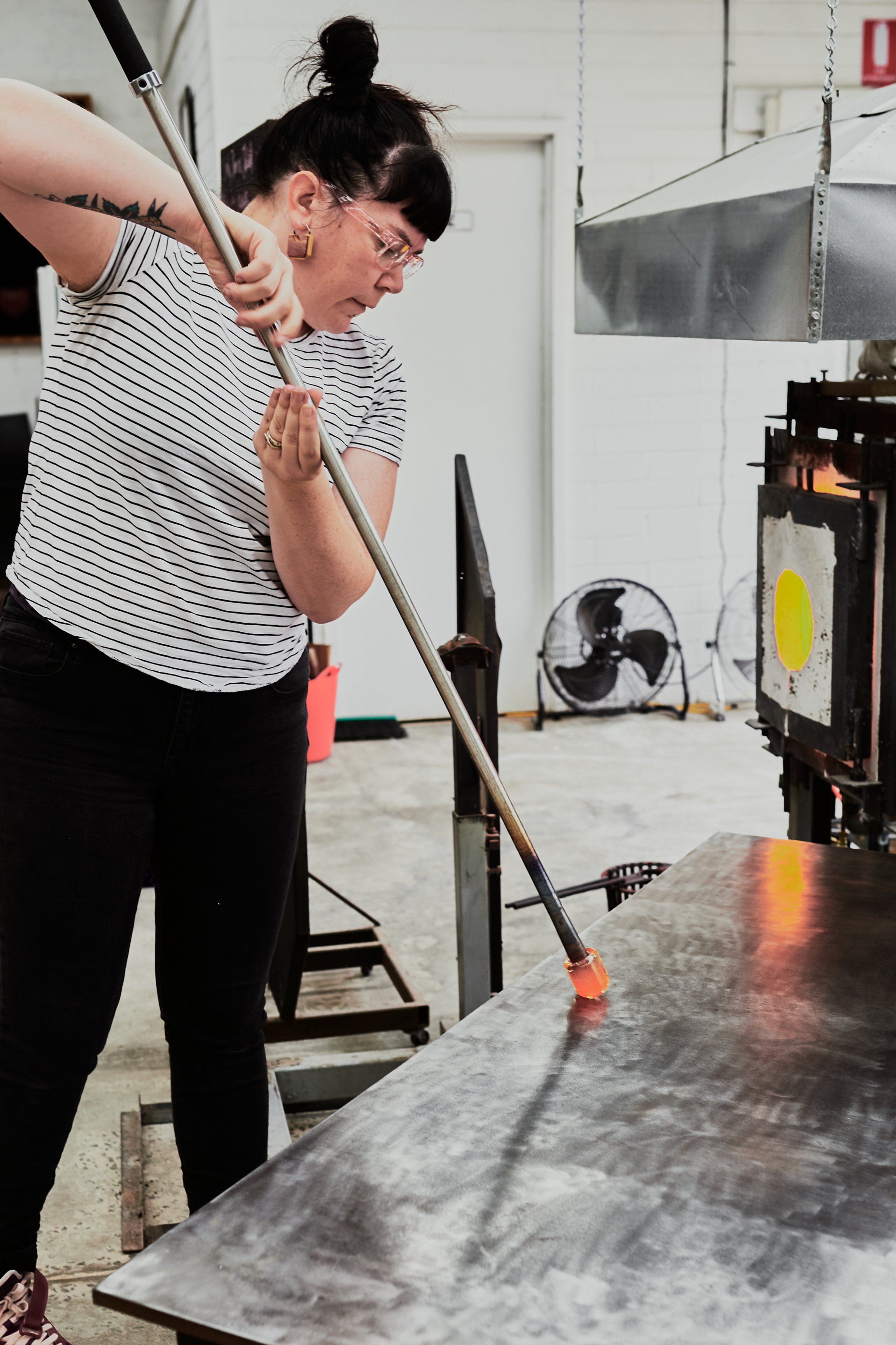 A woman wearing a striped black and white t-shirt holds a blow tube with hot glass on the end. She is pressing the hot glass onto a metal bench top and there is a furnace in the background.