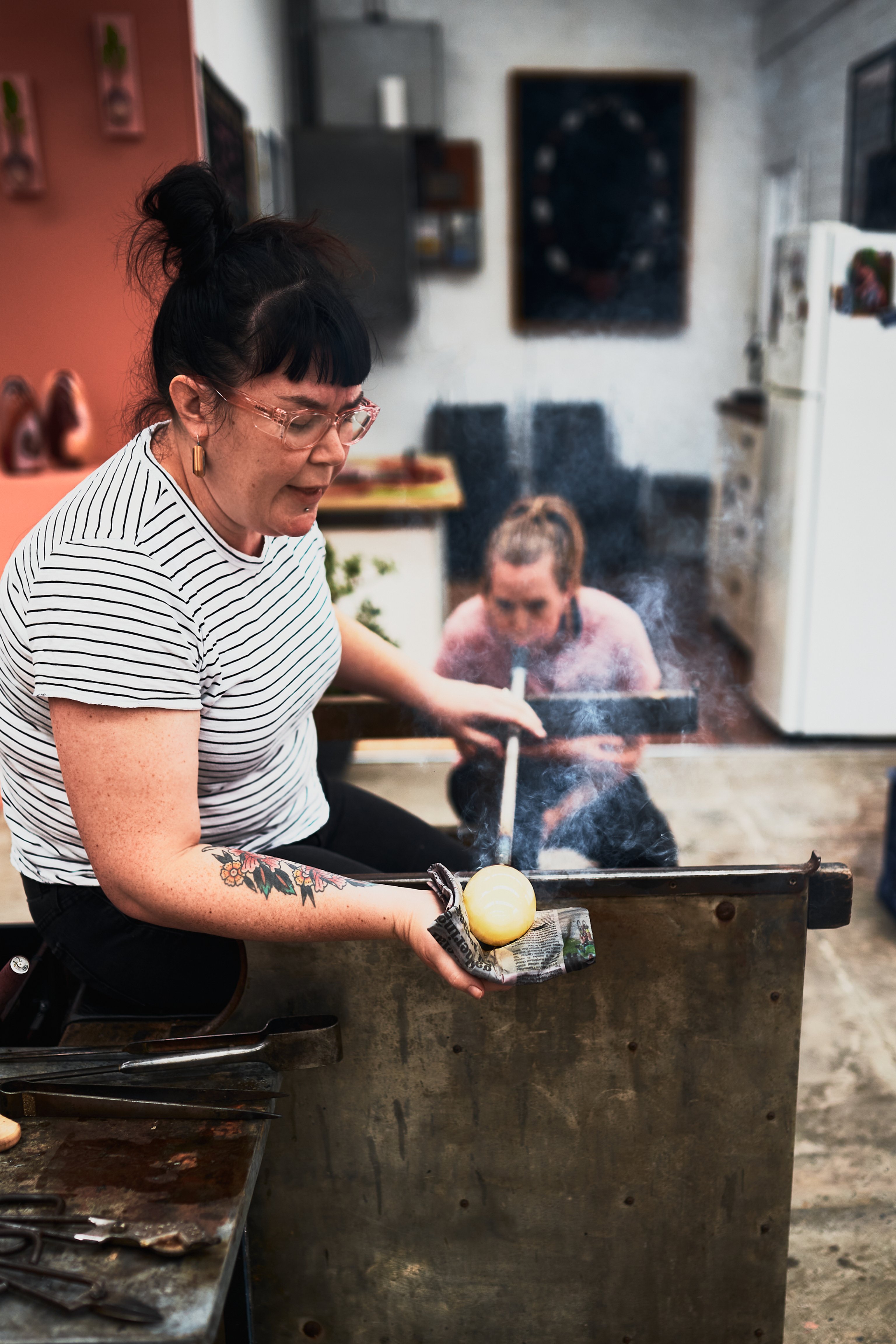 Two women are blowing glass in a workshop. One blows air into a long tube and the other cups the glass on the other end with wet newspaper.