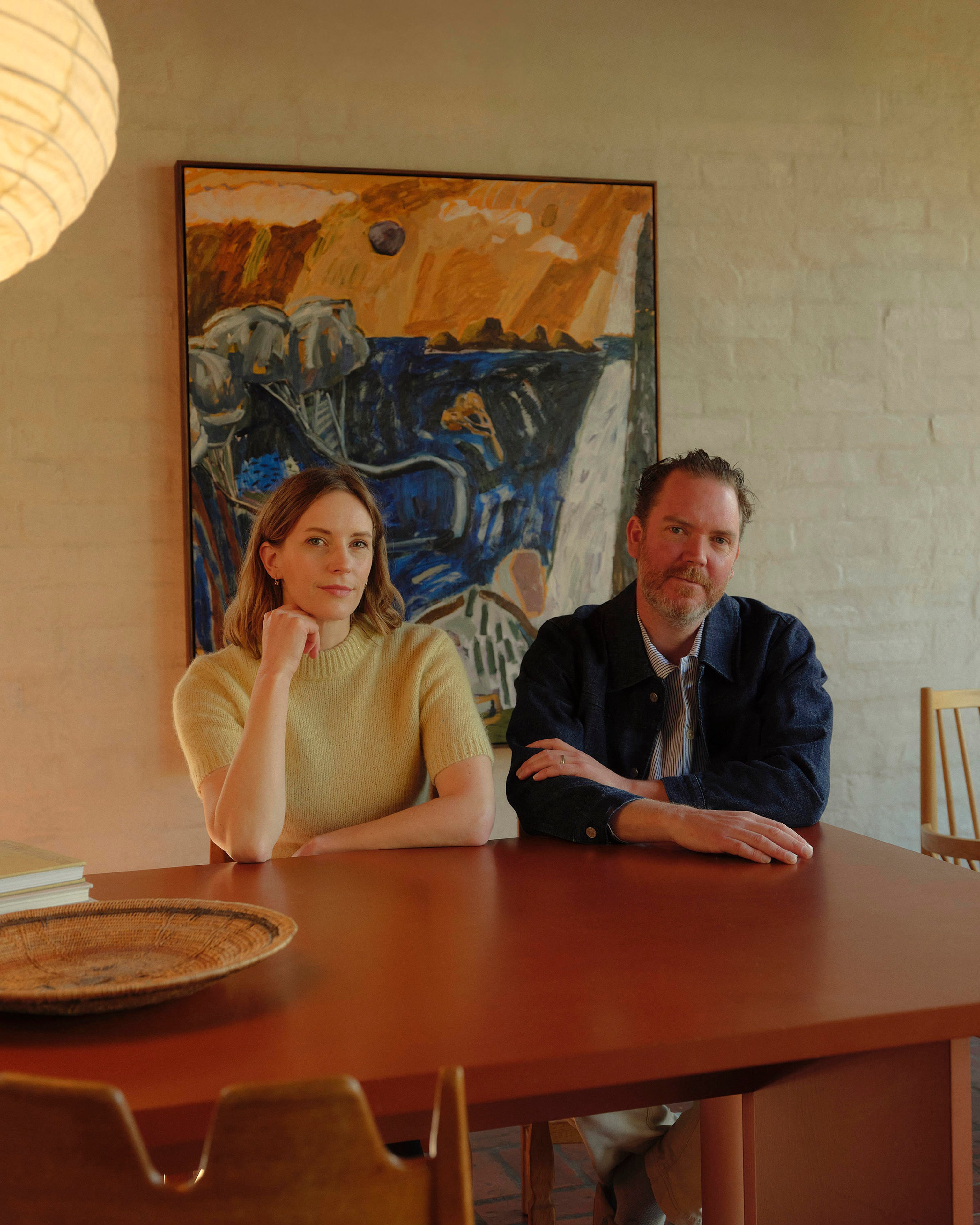 A portrait of Jess Lillico and Sean Fennessy, co-authors of Bush Modern, sitting at a dining table. On the rendered brick wall behind there is an abstract artwork.