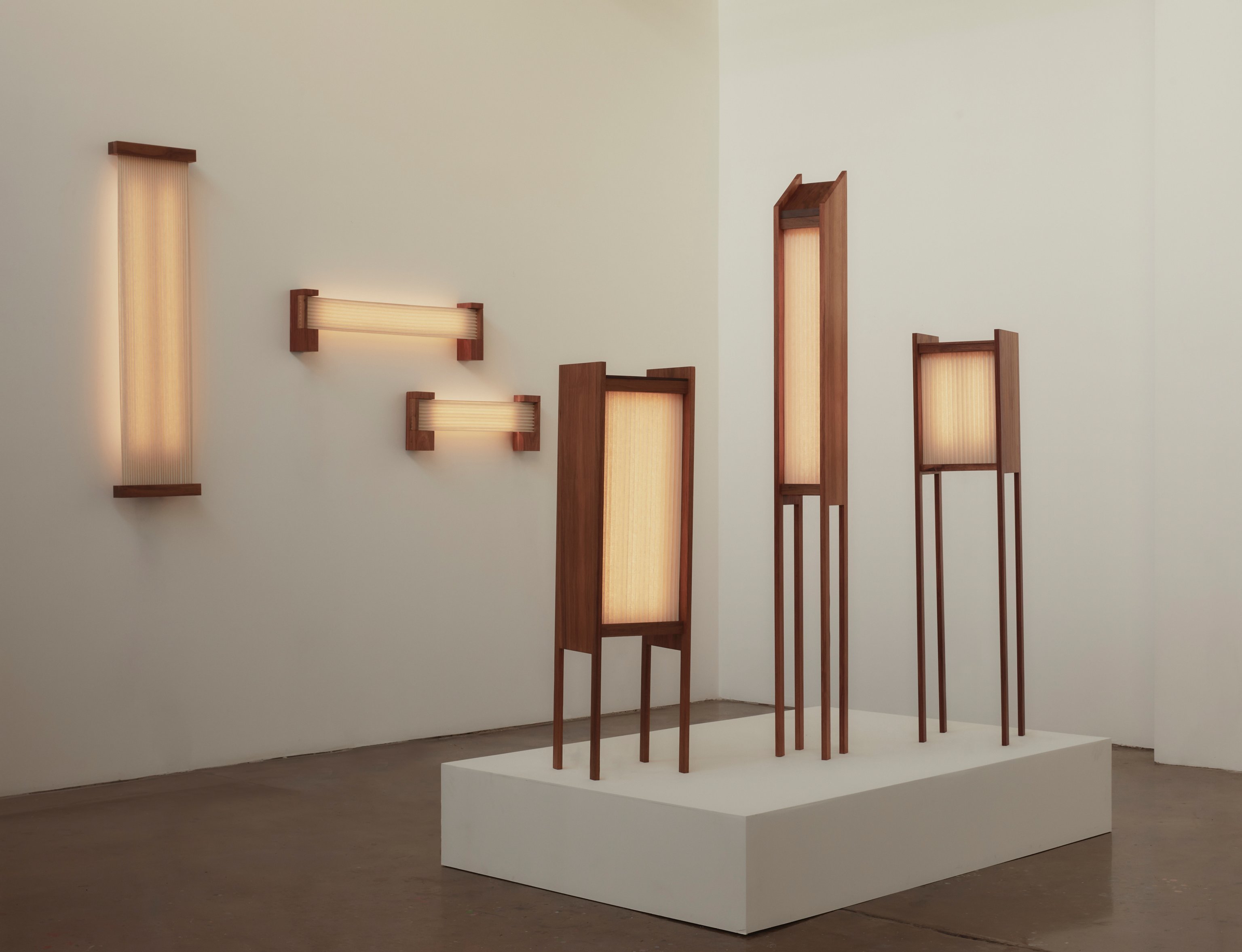 three floor lamps on a plinth and three wall lights hanging on the wall behind. All made with a timber frame and pleated linen.