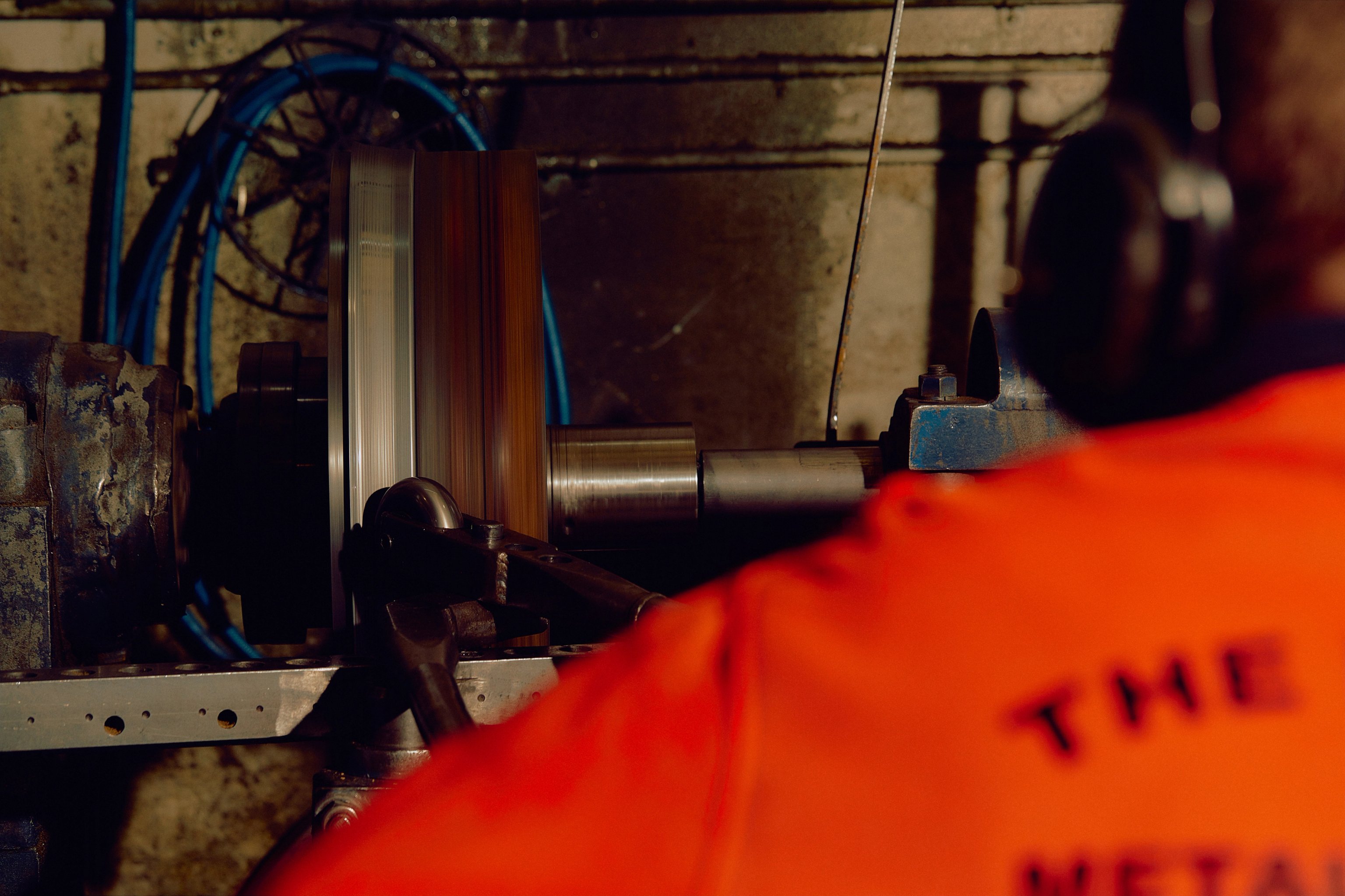 A man in a high visibility orange top is using a roller to spin metal on a lathe in an industrial factory.
