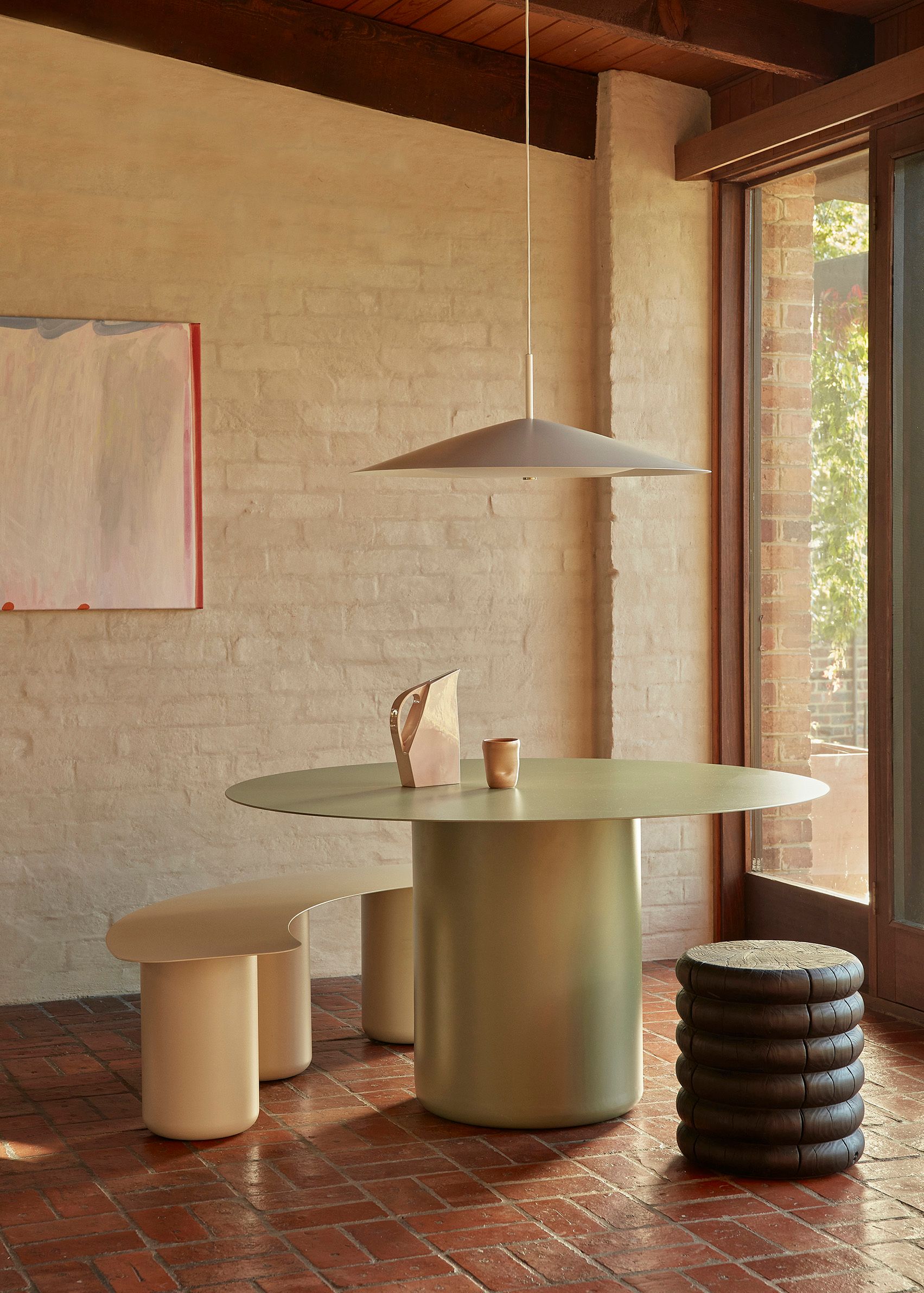 A pale eucalypt powder-coated steel dining table sits in a dining room with brick floors. The table is surrounded by a beige steel bench and wooden stool. A modern white pendant hangs from the ceiling