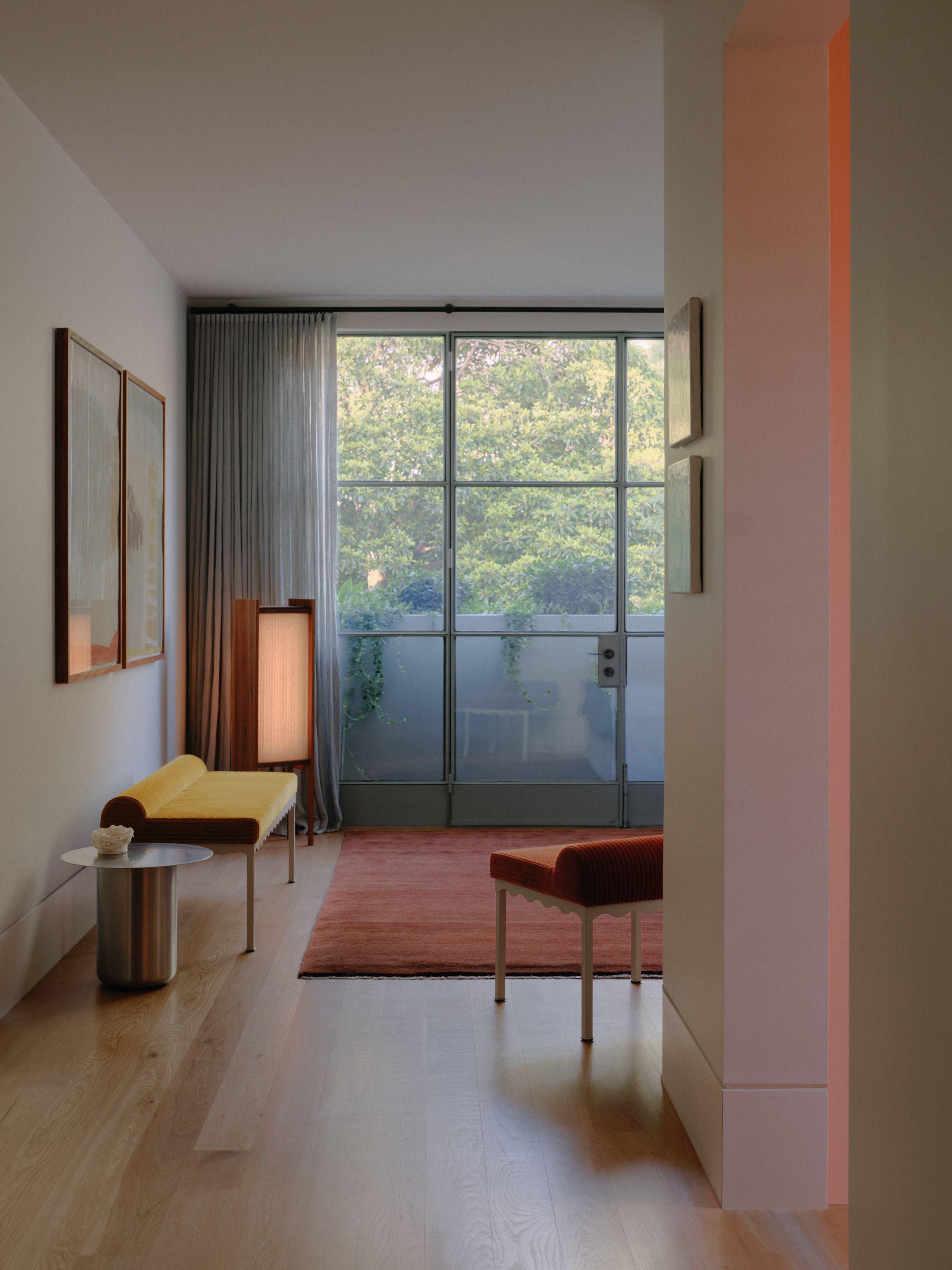 A modern room with large glass windows, lit up by a Linear pleated floor lamp. Also in the room is a rich terracotta rug, red and yellow upholstered benches and a stainless steel side table
