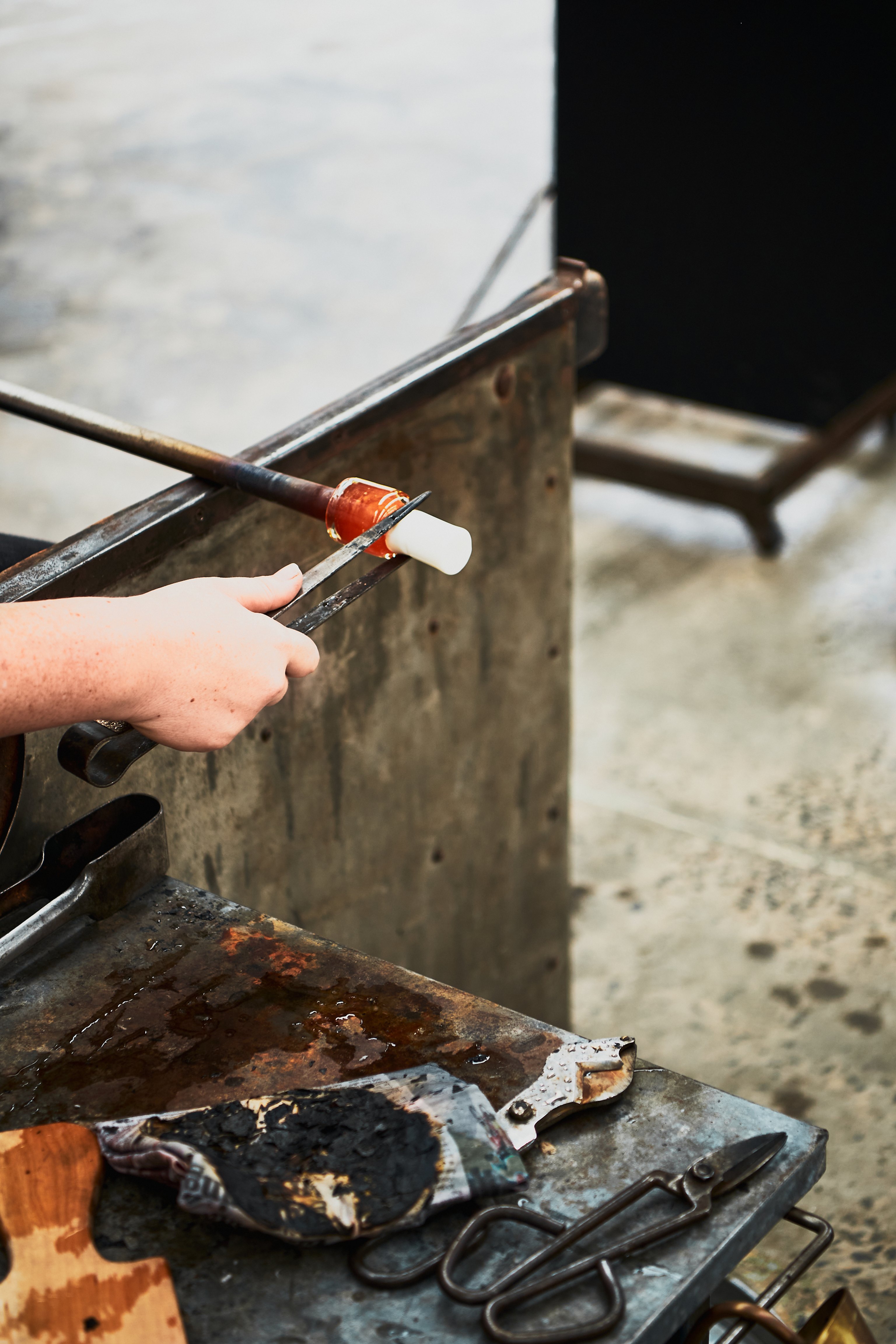 A hand hold a pair of tongs that clamp a small rod of white glass on the end of a blow tube. There are some metal tools on a metal workbench in the foreground.