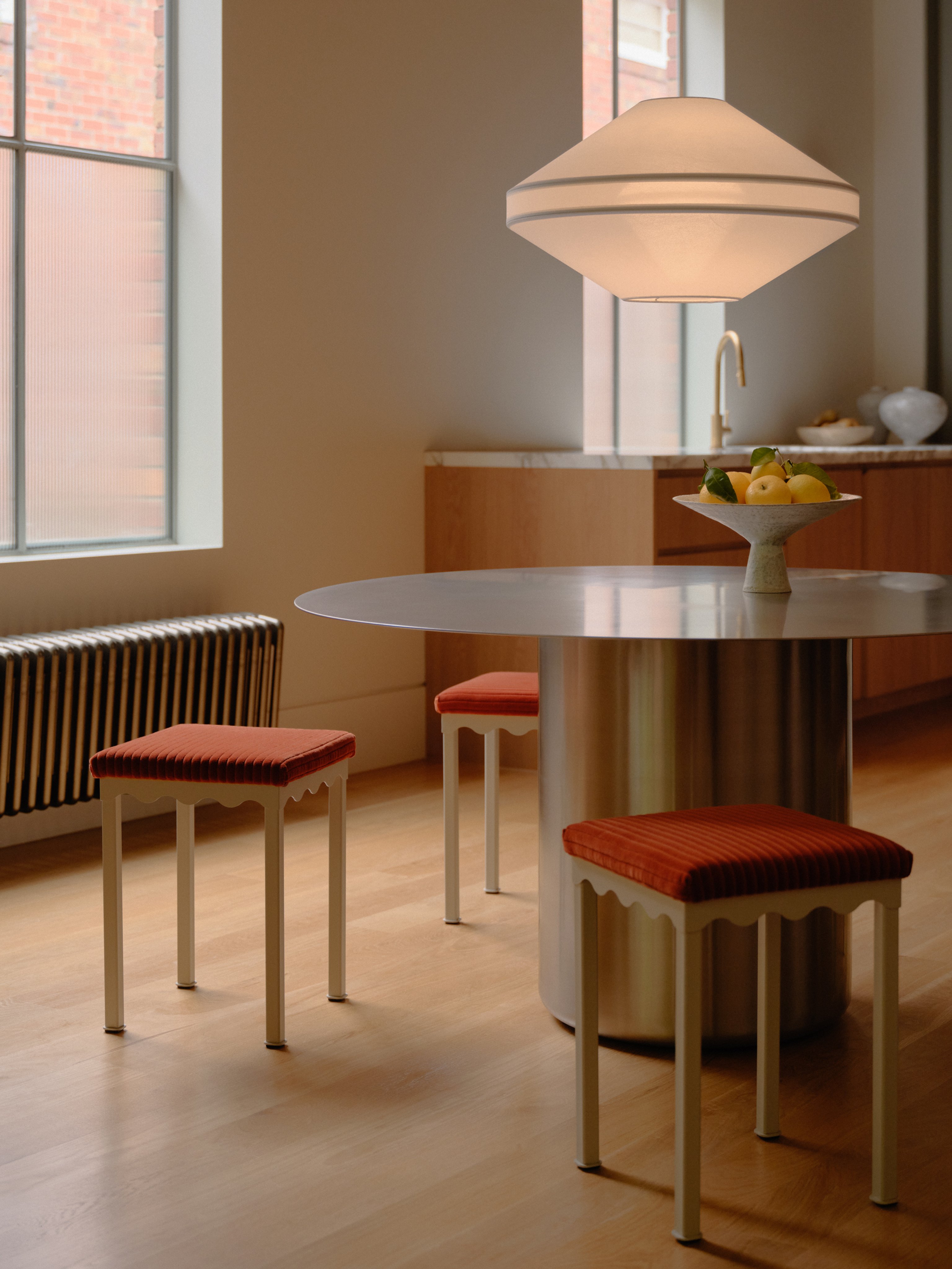 A large mesh fabric pendant light in an oval shape sits above a stainless steel dining table with red corduroy stools