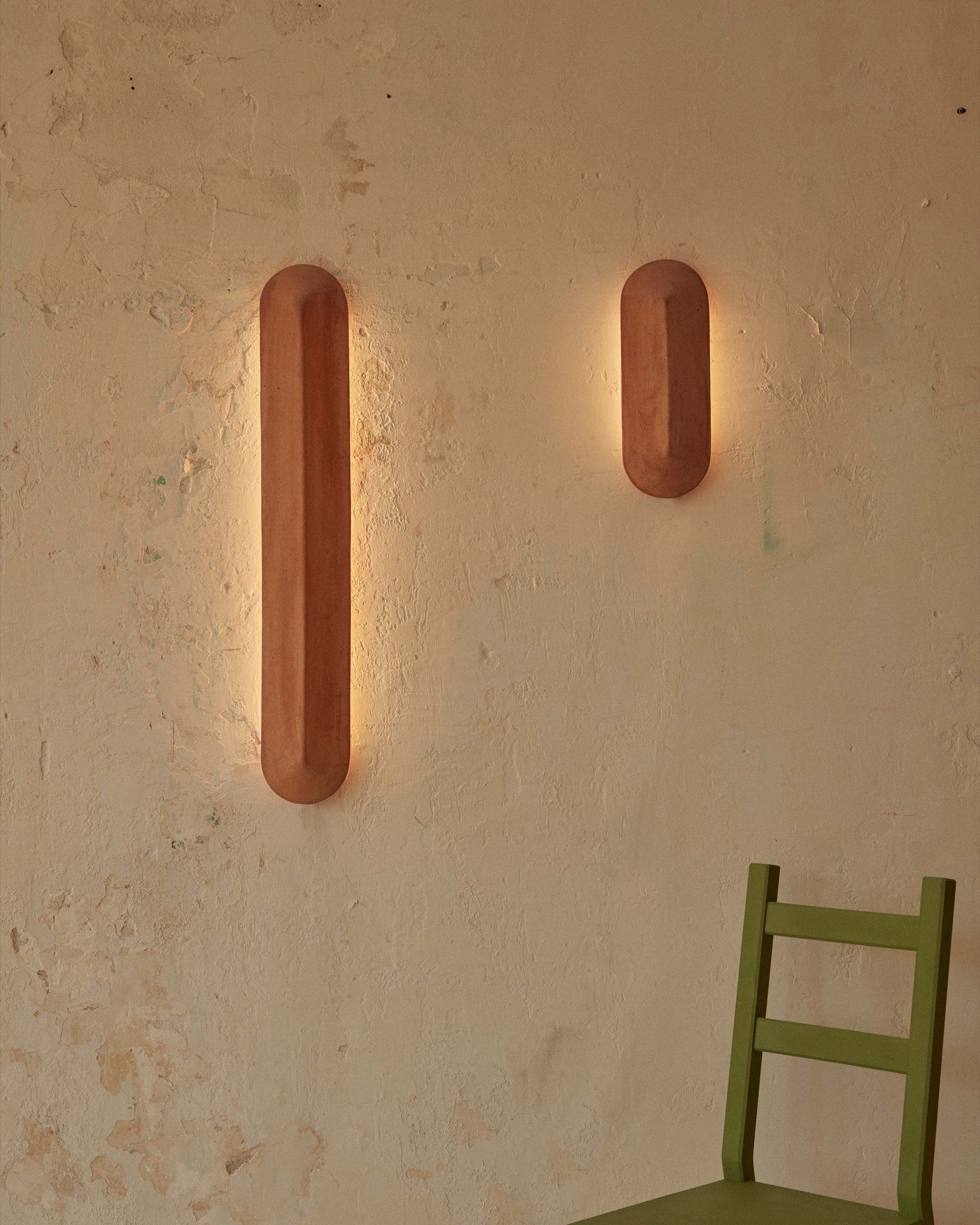 Two terracotta Pinch wall lights fixed to a cream wall illuminating a green timber chair
