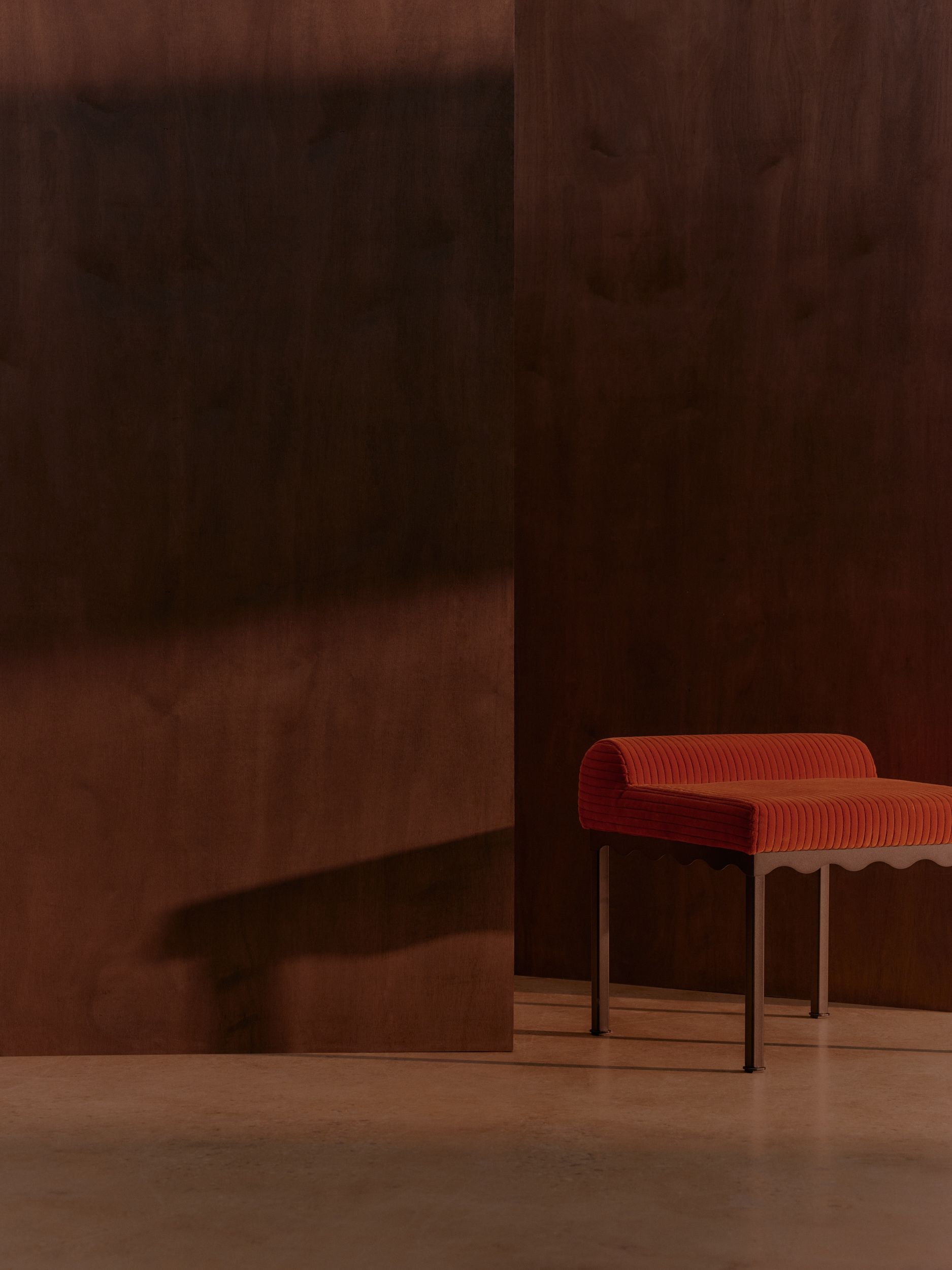 A red corduroy upholstered bench seat featuring wavy black metal frames in a timber room with dim lighting and strong shadows
