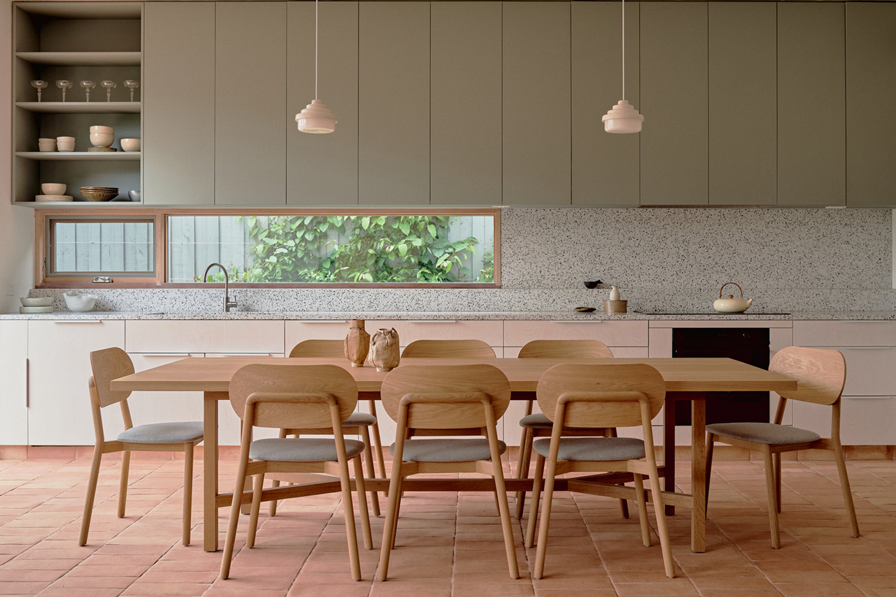 A large kitchen and dining area with green cabinetry, ceramic pendant lights and timber table setting
