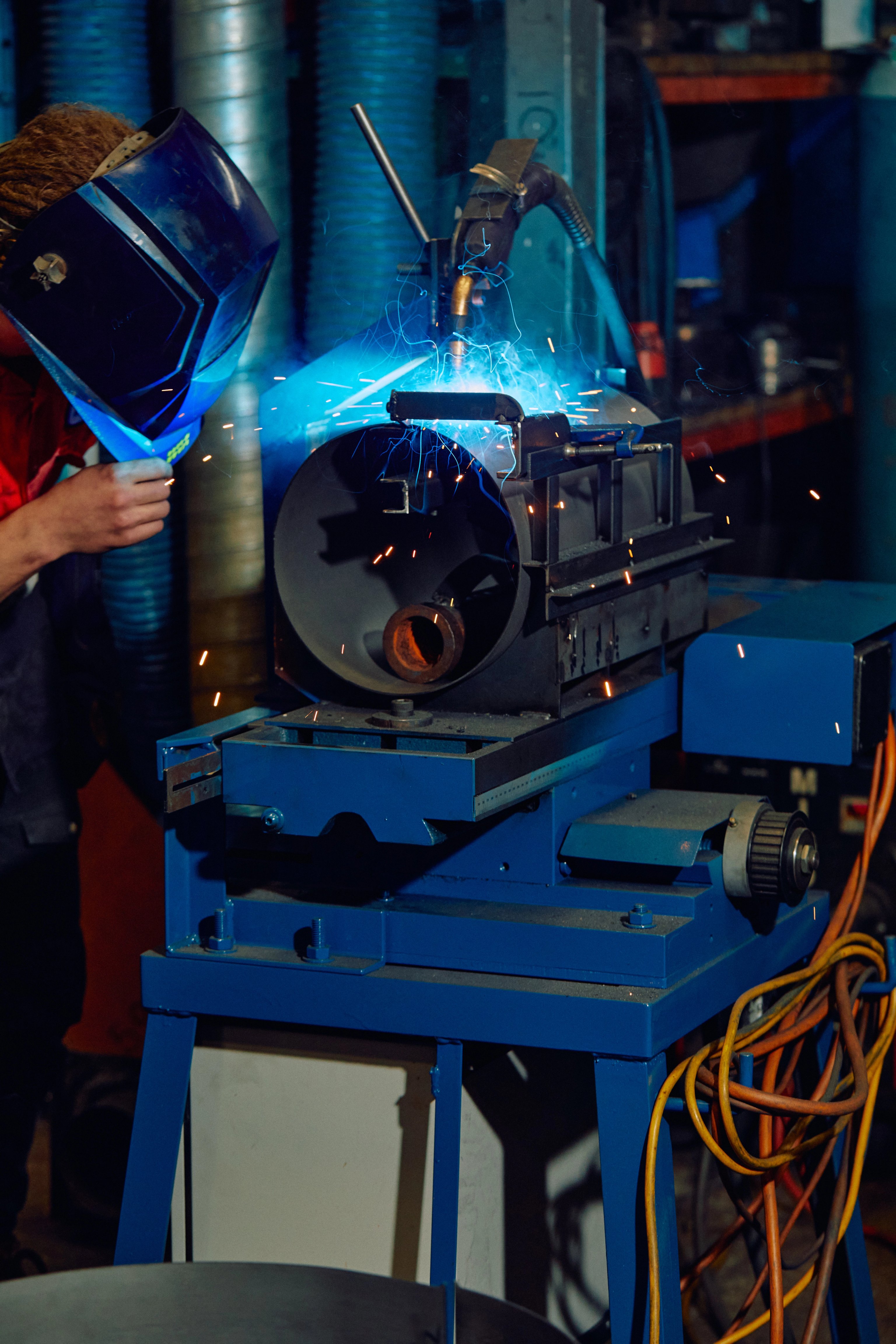 A metal drum being welded by a man wearing a safety mast. Blue sparks are flying around.