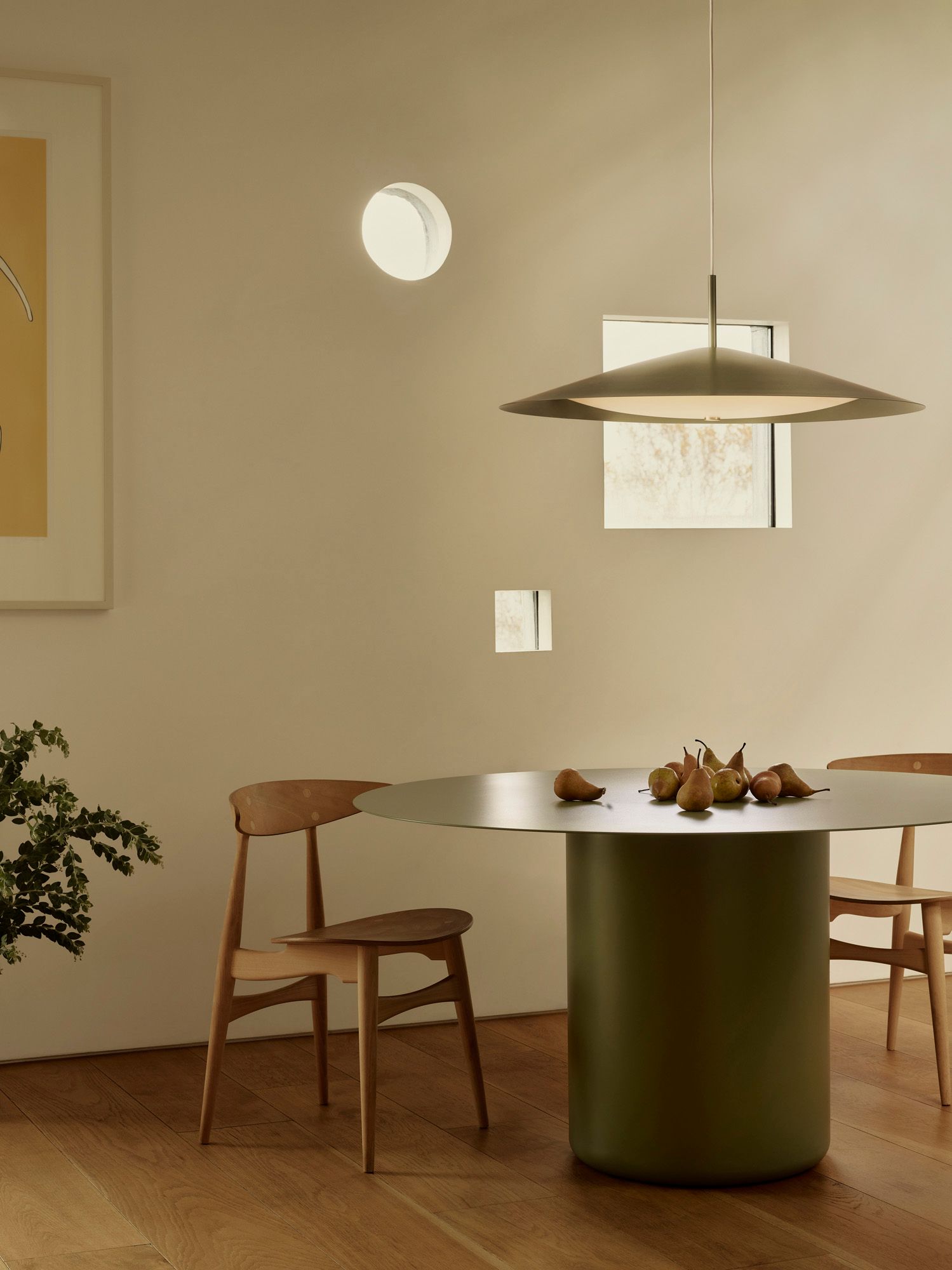 A large green pendant light is placed above a green round dining table. The table is set with two wooden dining chairs