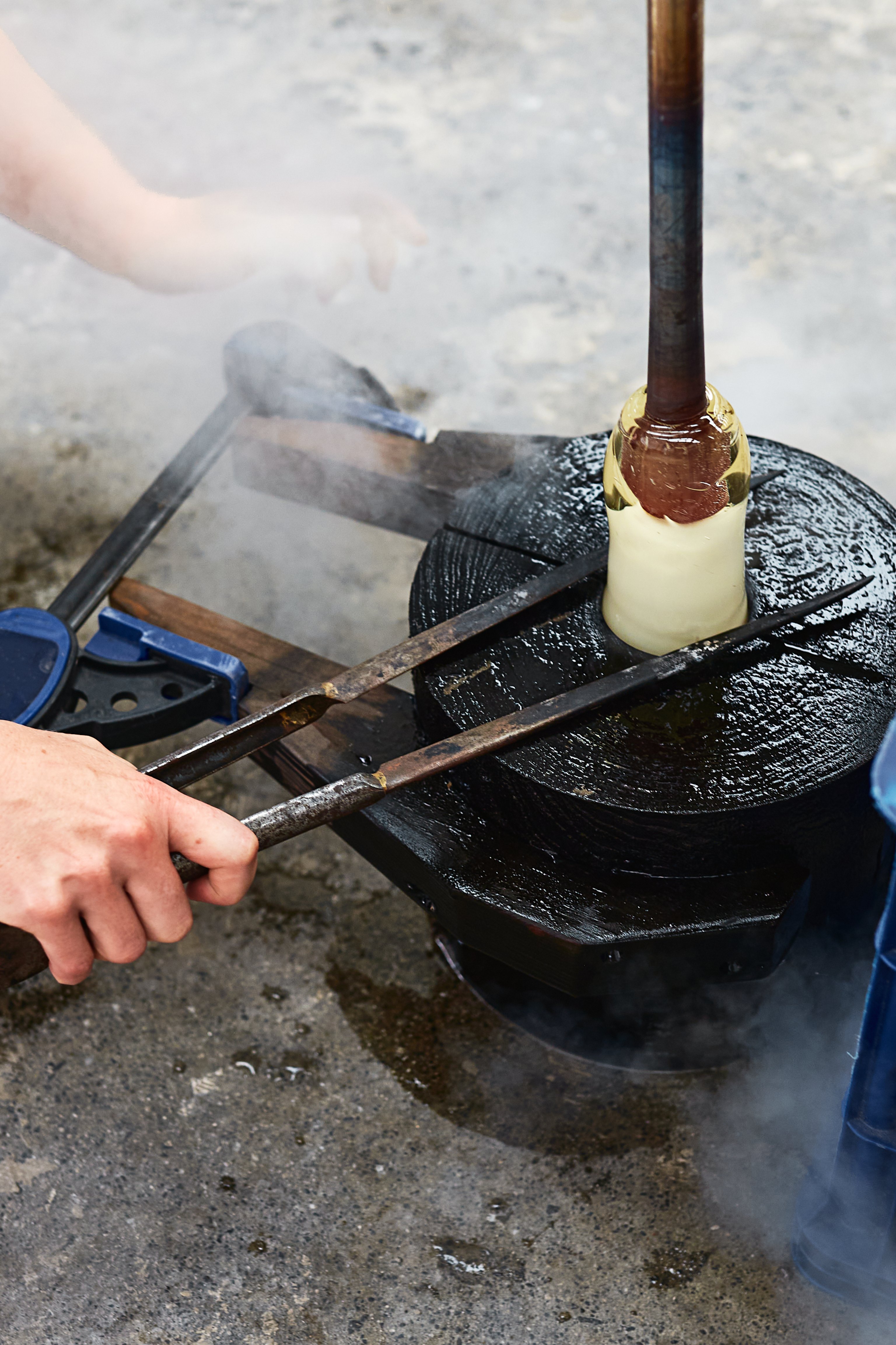 A pair of hands use tongs to clamp hot glass inside a timber glass blowing mould. Steam is pouring out of the mould due to the heat.