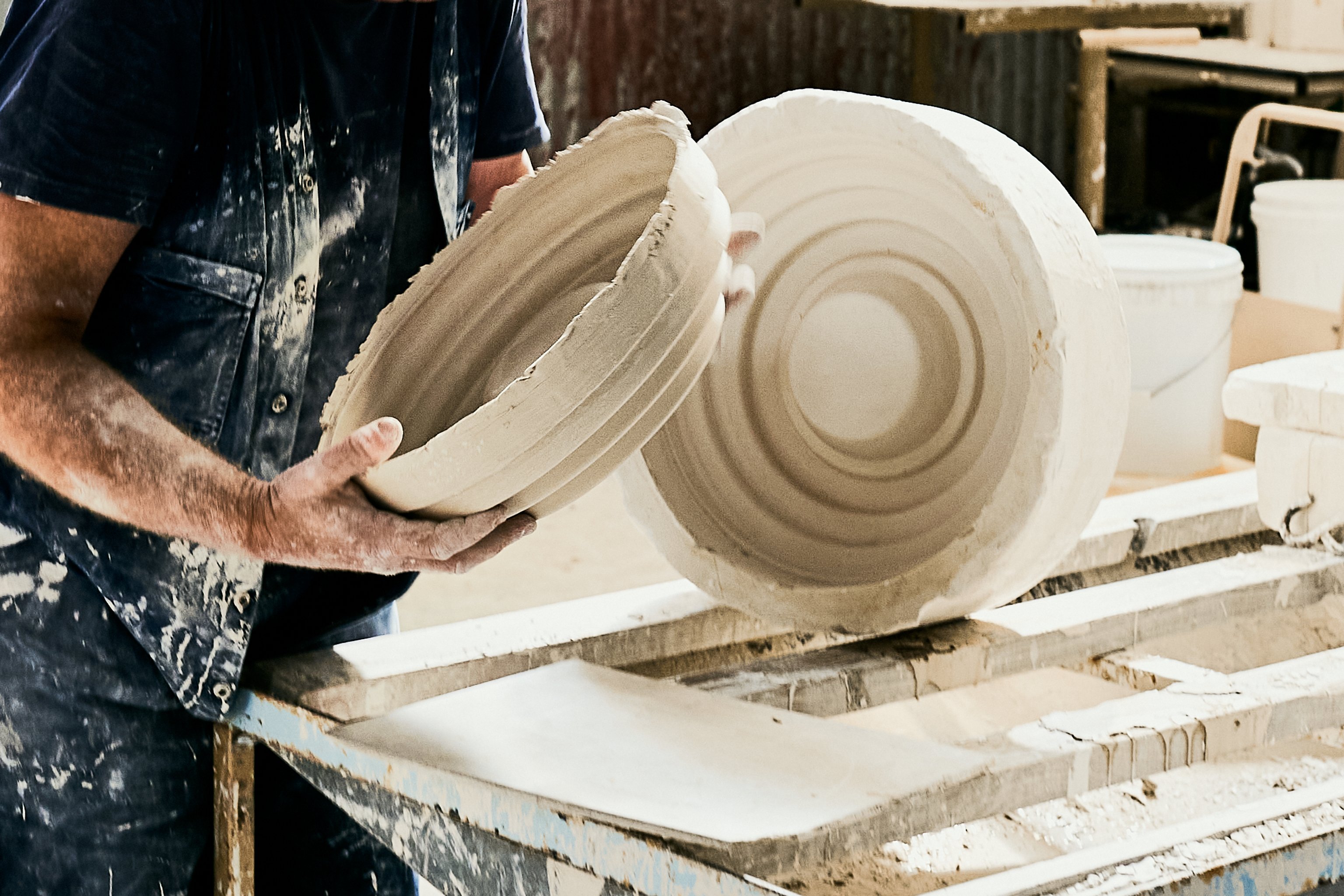 A man holds a ceramic 2-part mould for the Honey Wall Lights open. He is wearing overalls covered in clay.