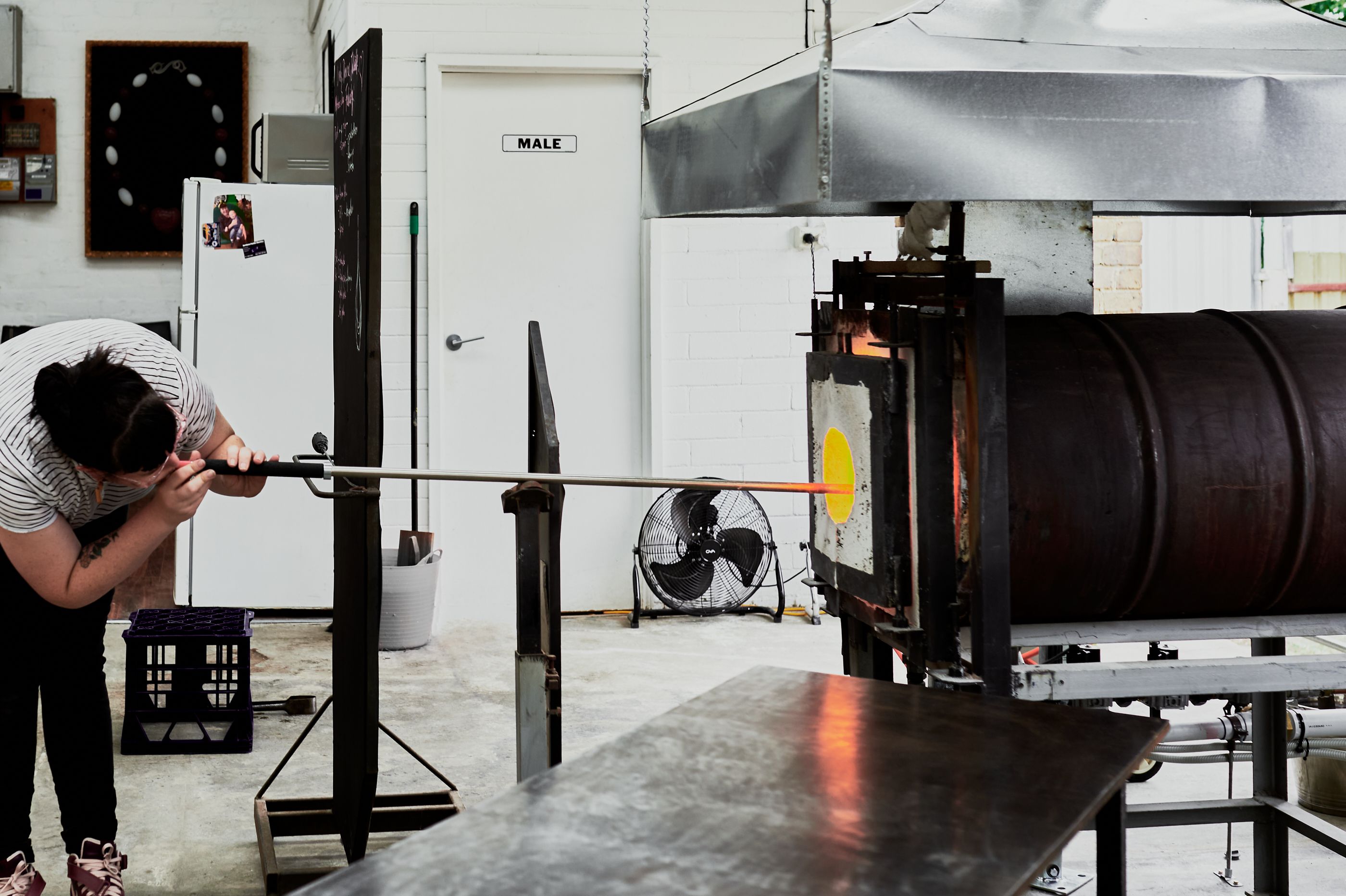 A woman is blowing glass, she bends down and blows air into a long tube that enters into the furnace. 