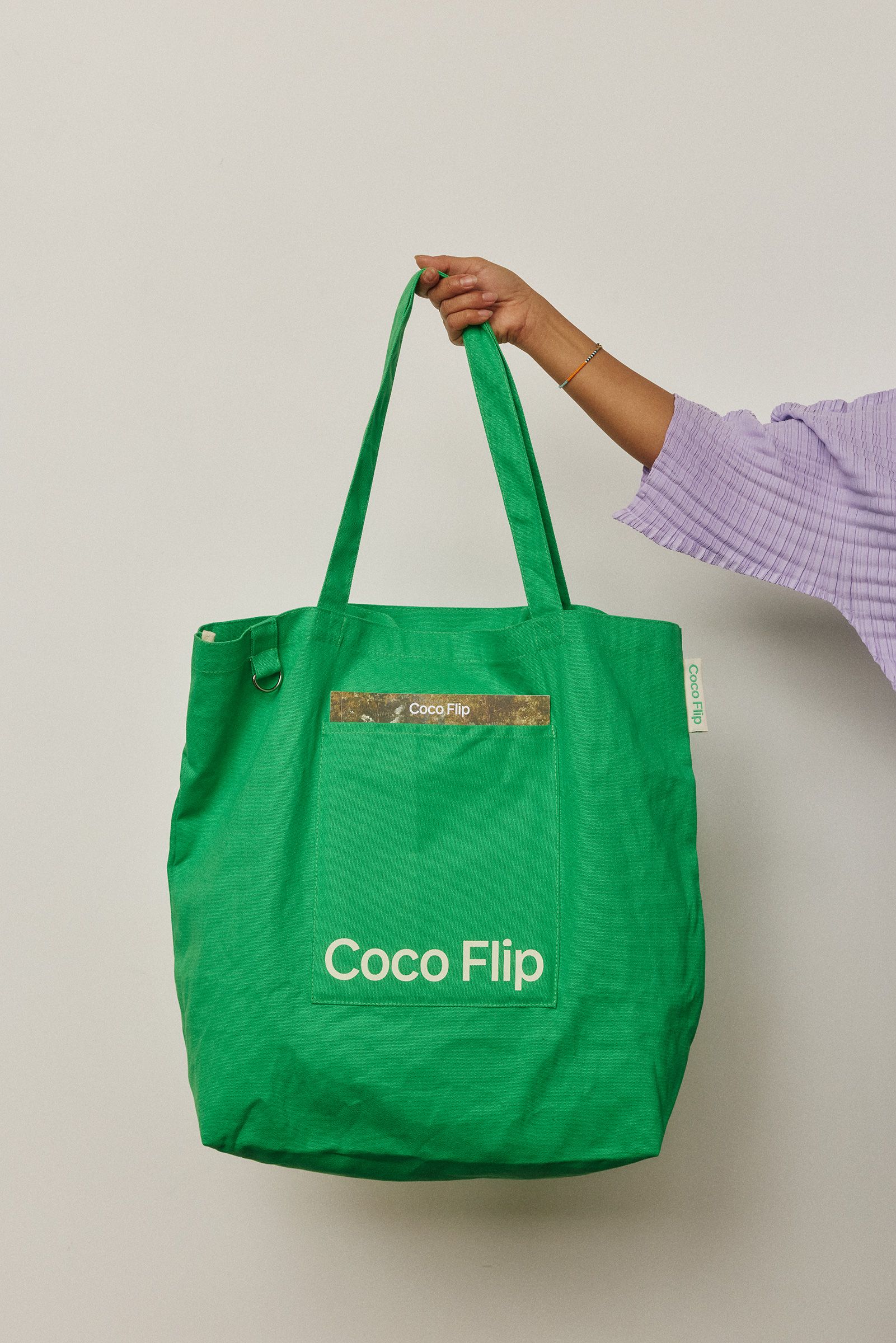 A green tote bag with Coco Flip logo being held by a woman in a purple top
