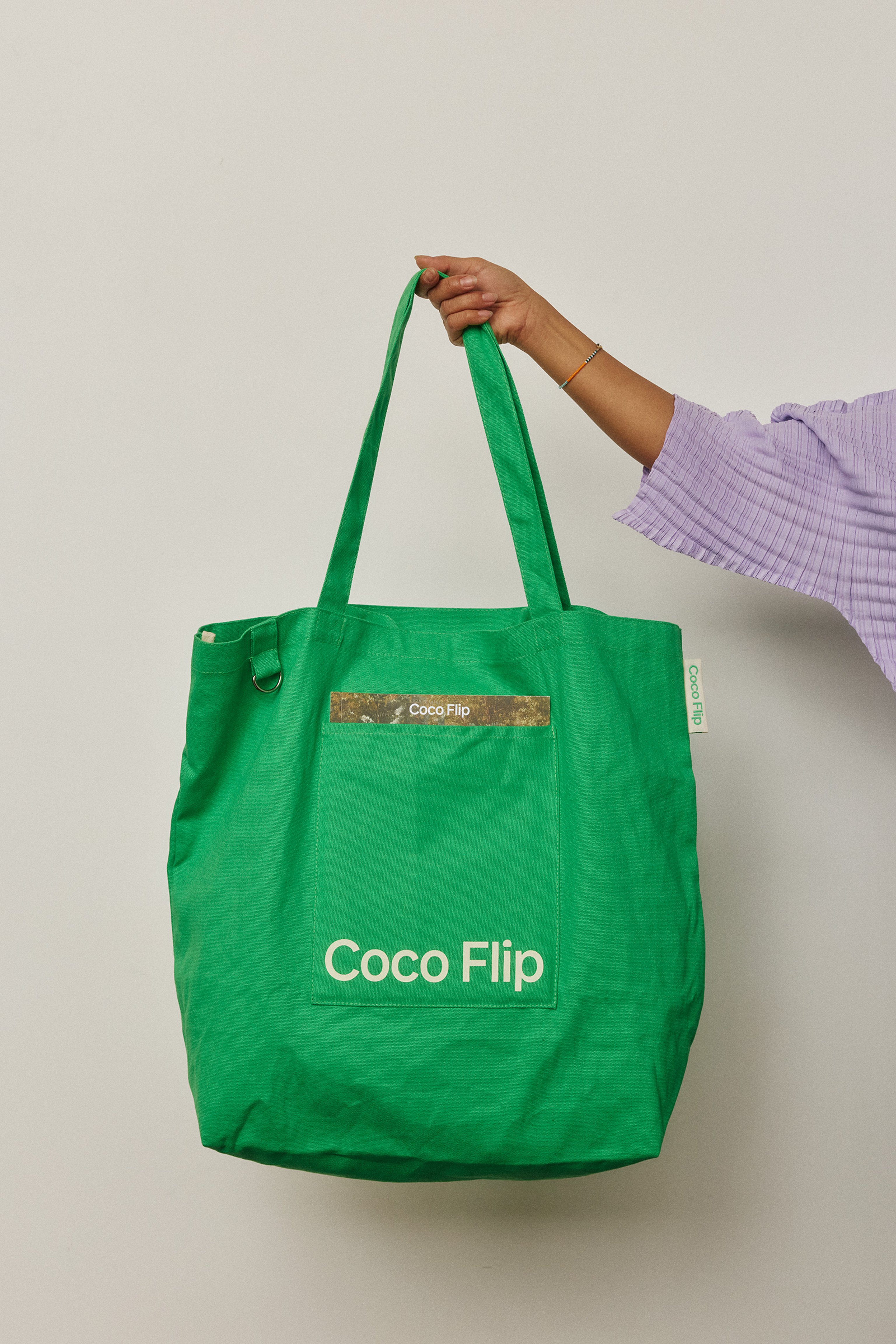 A green tote bag with Coco Flip logo being held by a woman in a purple top