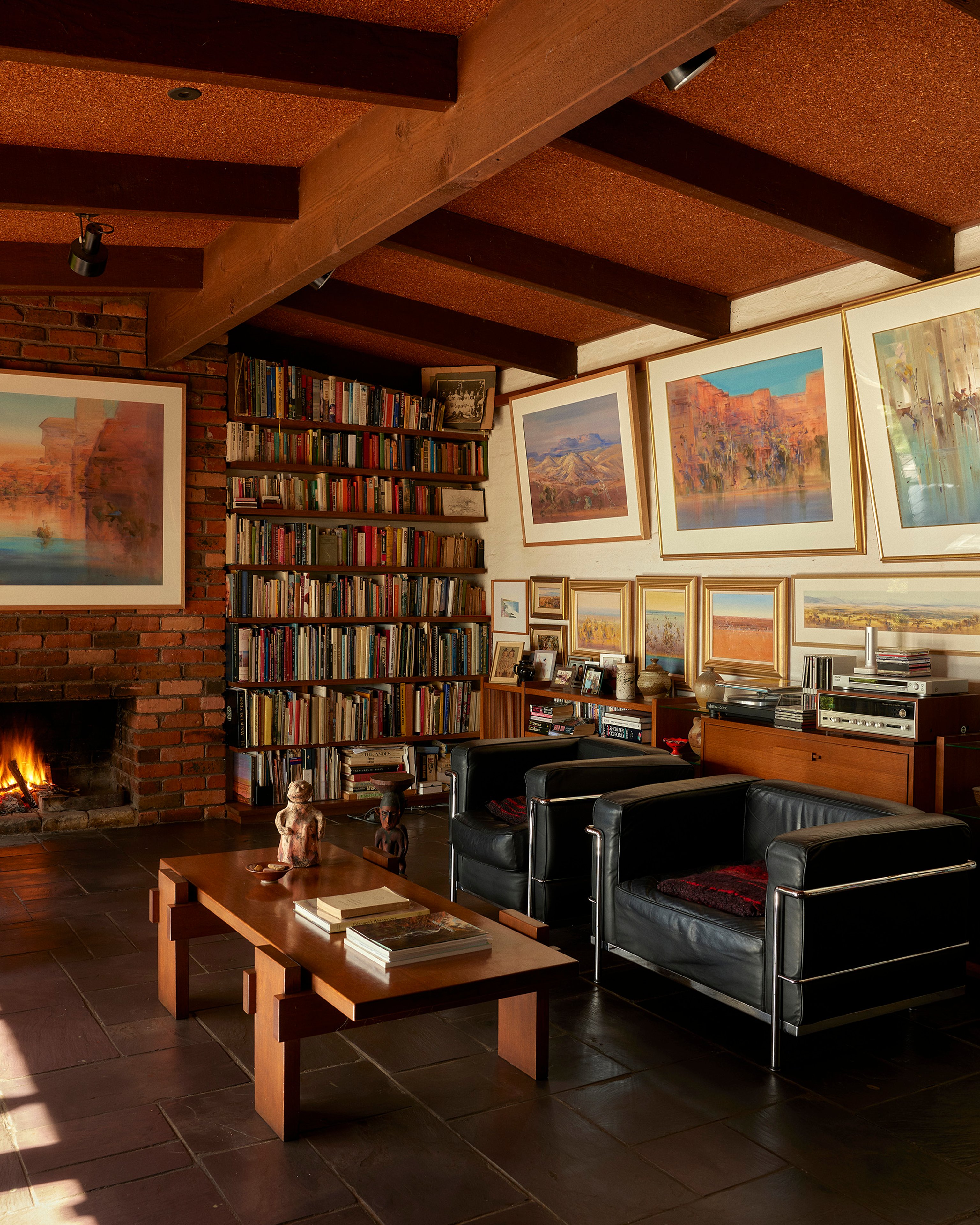 A richly textured modernist home interior with art filling the walls and books filling the bookshelves. Two black leather armchairs and a timber coffee table furnish the room.