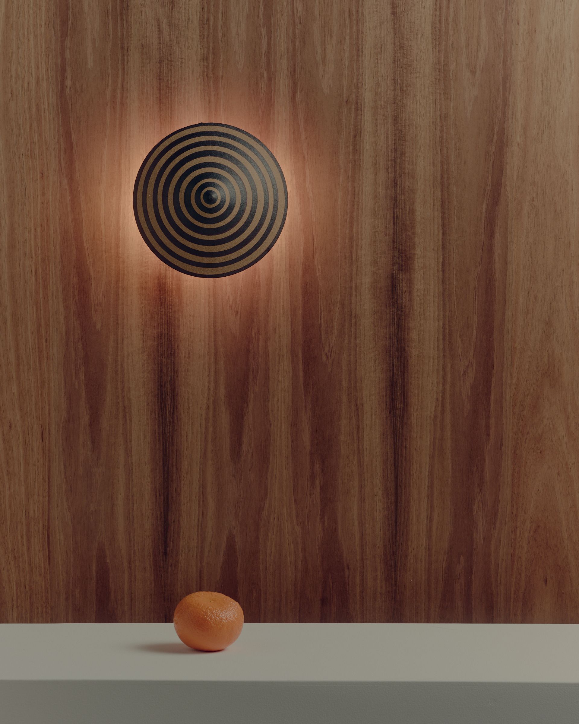 A round striped ceramic Dancer Wall Light mounted on a timber wall with an orange in the foreground