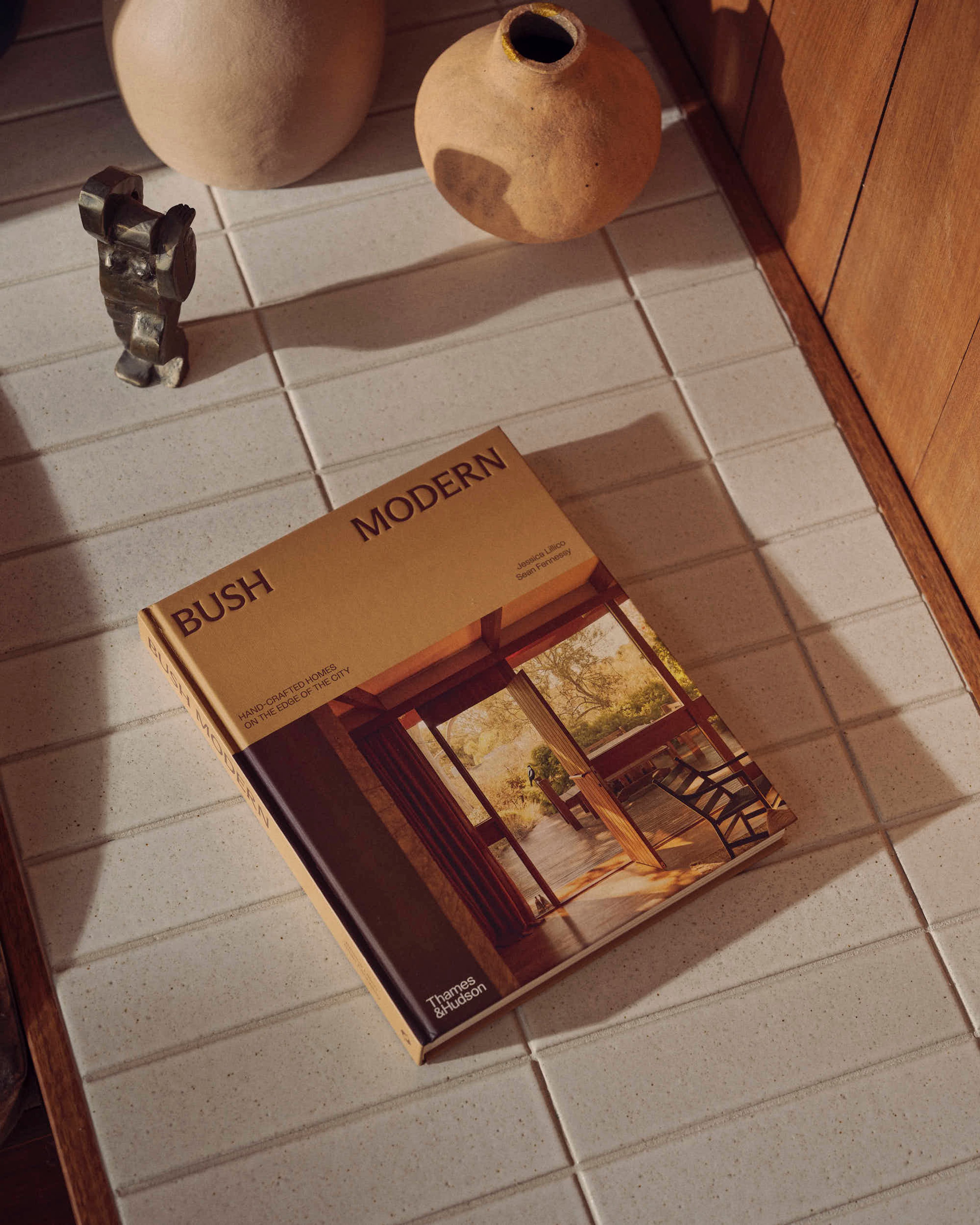 A copy of the book Bush Modern sits on a tiled surface in warm daylight with some vessels and a sculpture also visable, shot from above.