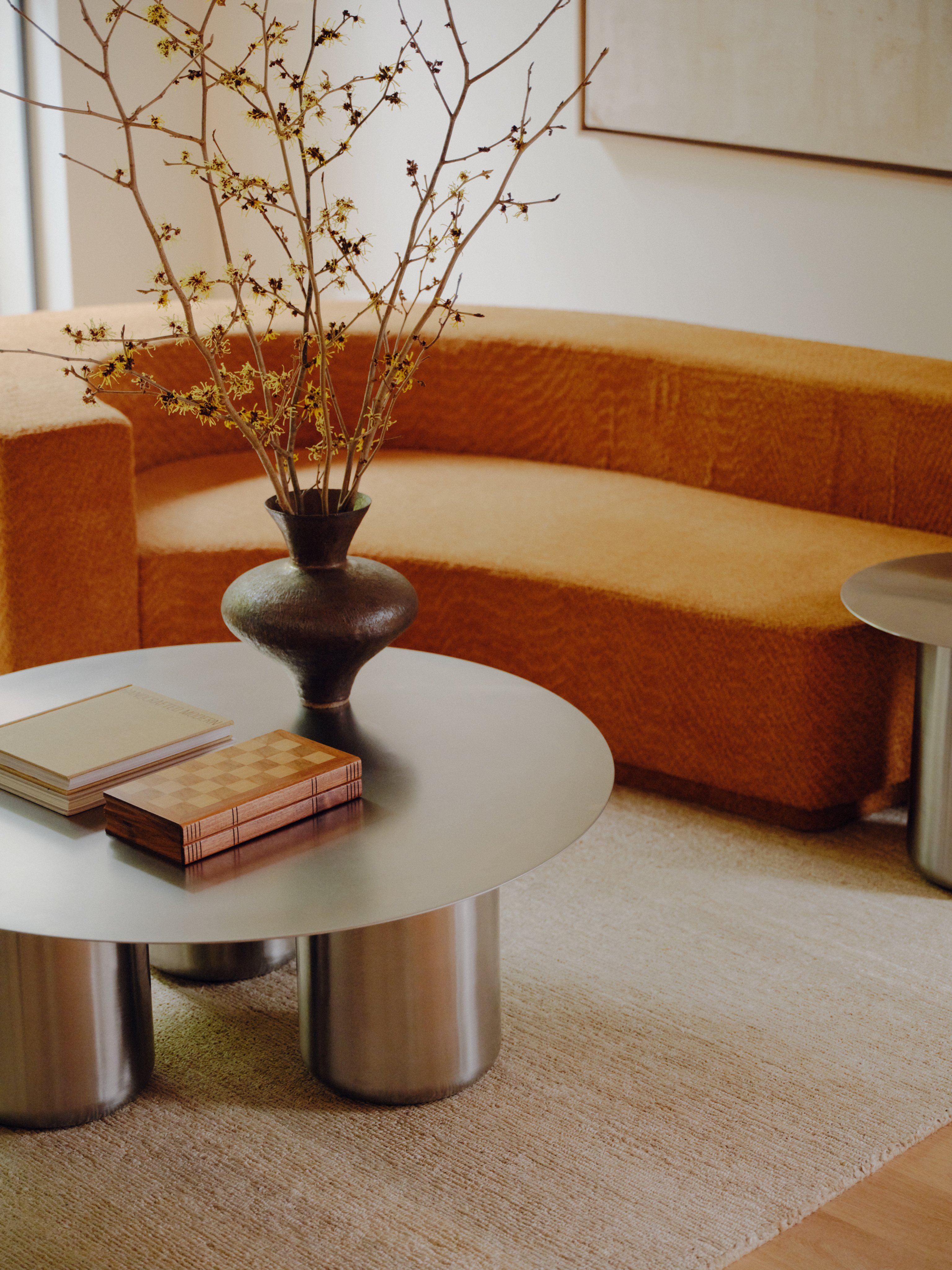 A Sequence Round Coffee Table in stainless steel, on a light coloured rug next to a burnt orange sofa.