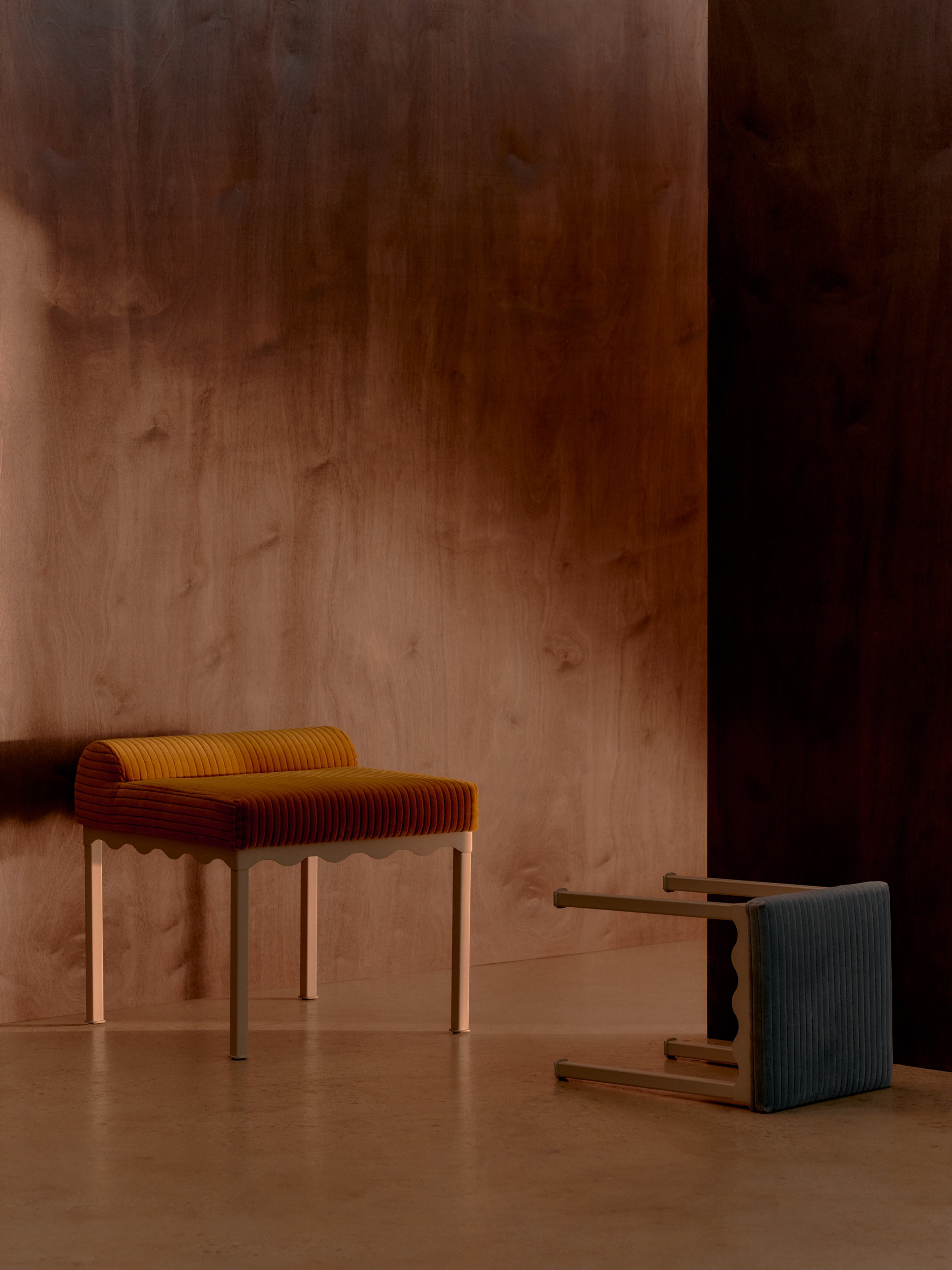 A yellow corduroy upholstered bench seat and blue corduroy upholstered stool featuring wavy metal frames in a timber room with dim lighting and strong shadows