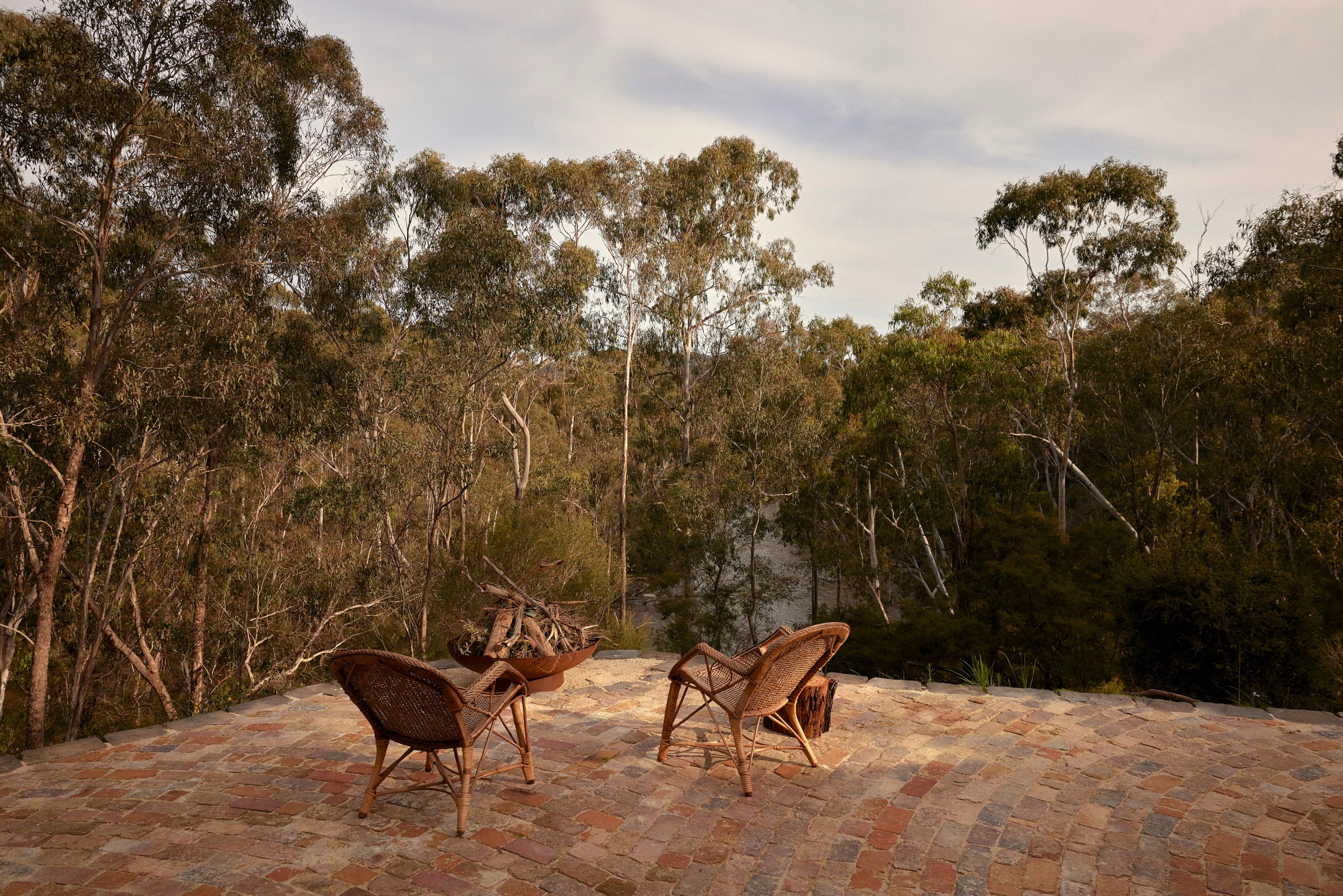 Two cane chairs and a rusted fireplace stacked with wood, sitting on brick paving and looking out over a river surrounded by large gum trees and bushland