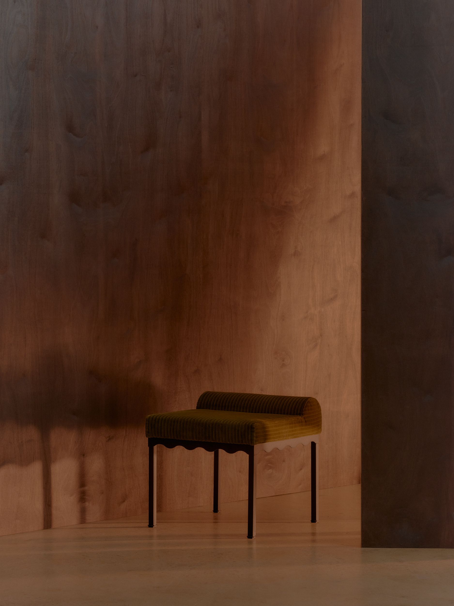 A green corduroy upholstered bench seat featuring wavy stainless steel frames in a timber room with dim lighting and strong shadows