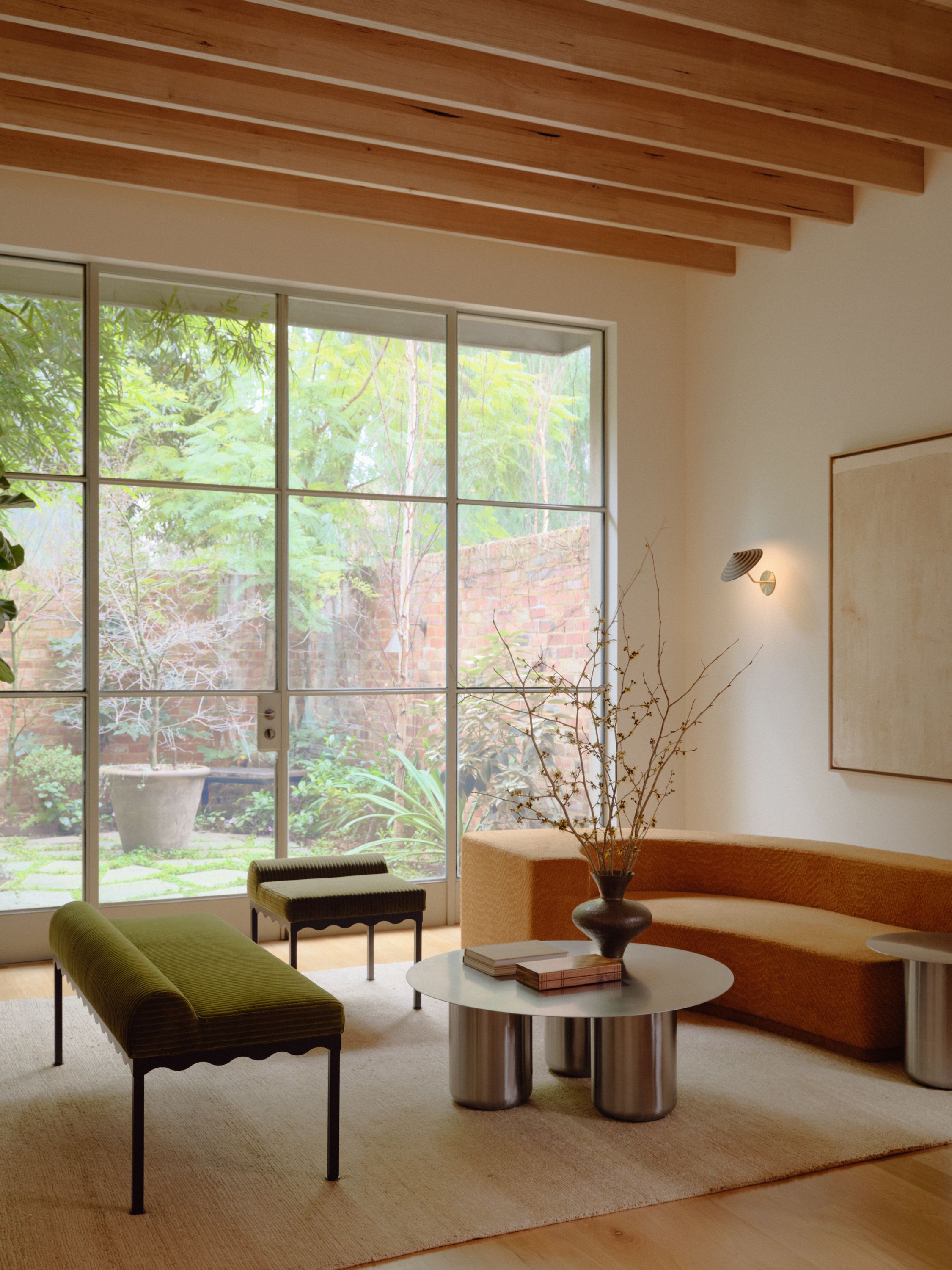 A living room setting with a ceramic striped Dancer Angled Wall Light and a Bellini Upholstered Bench. Windows look out onto greenery.