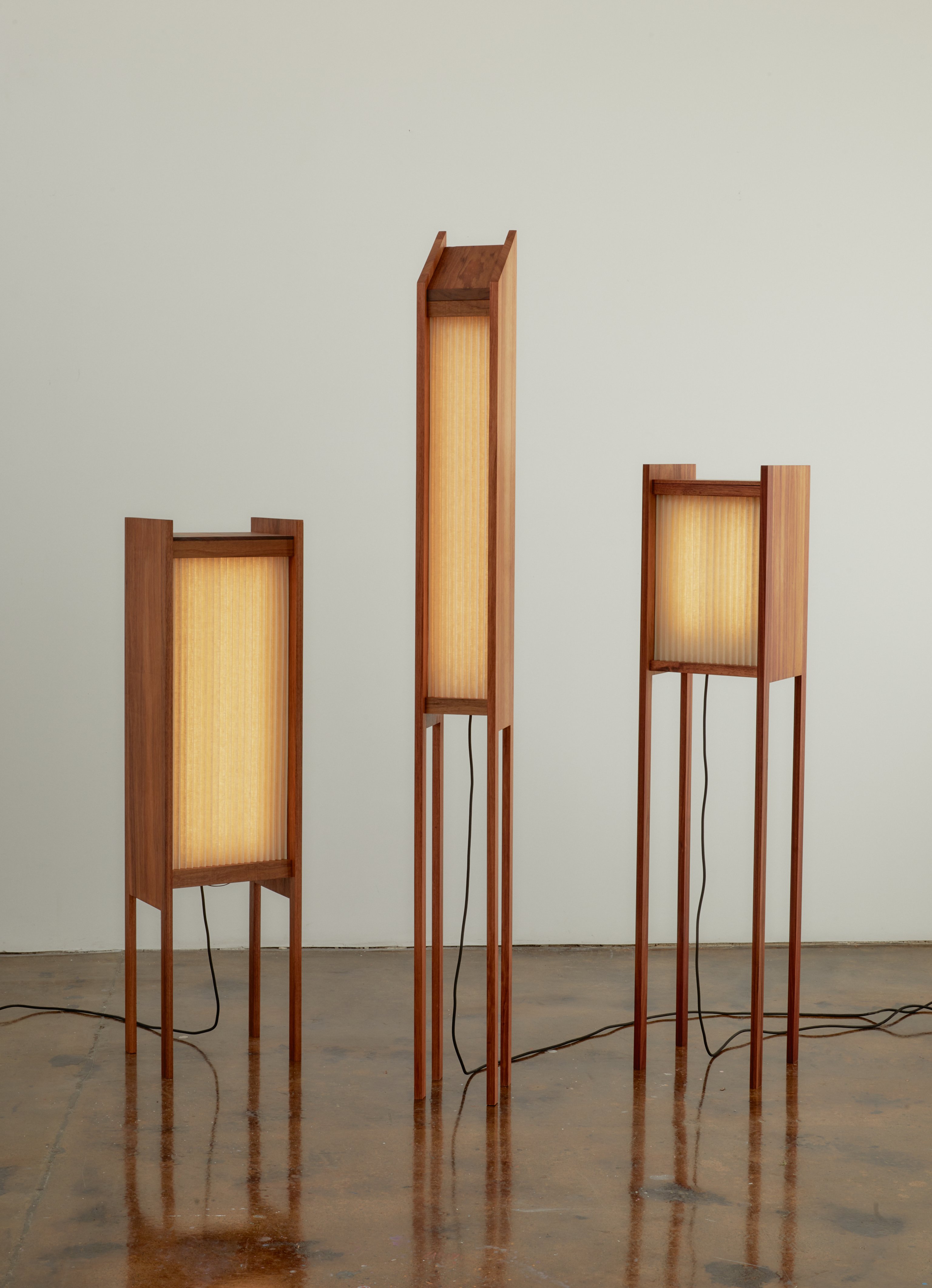 Three timber and pleated linen floor lamps with tall, skinny legs on a white background. They have a warm glow.