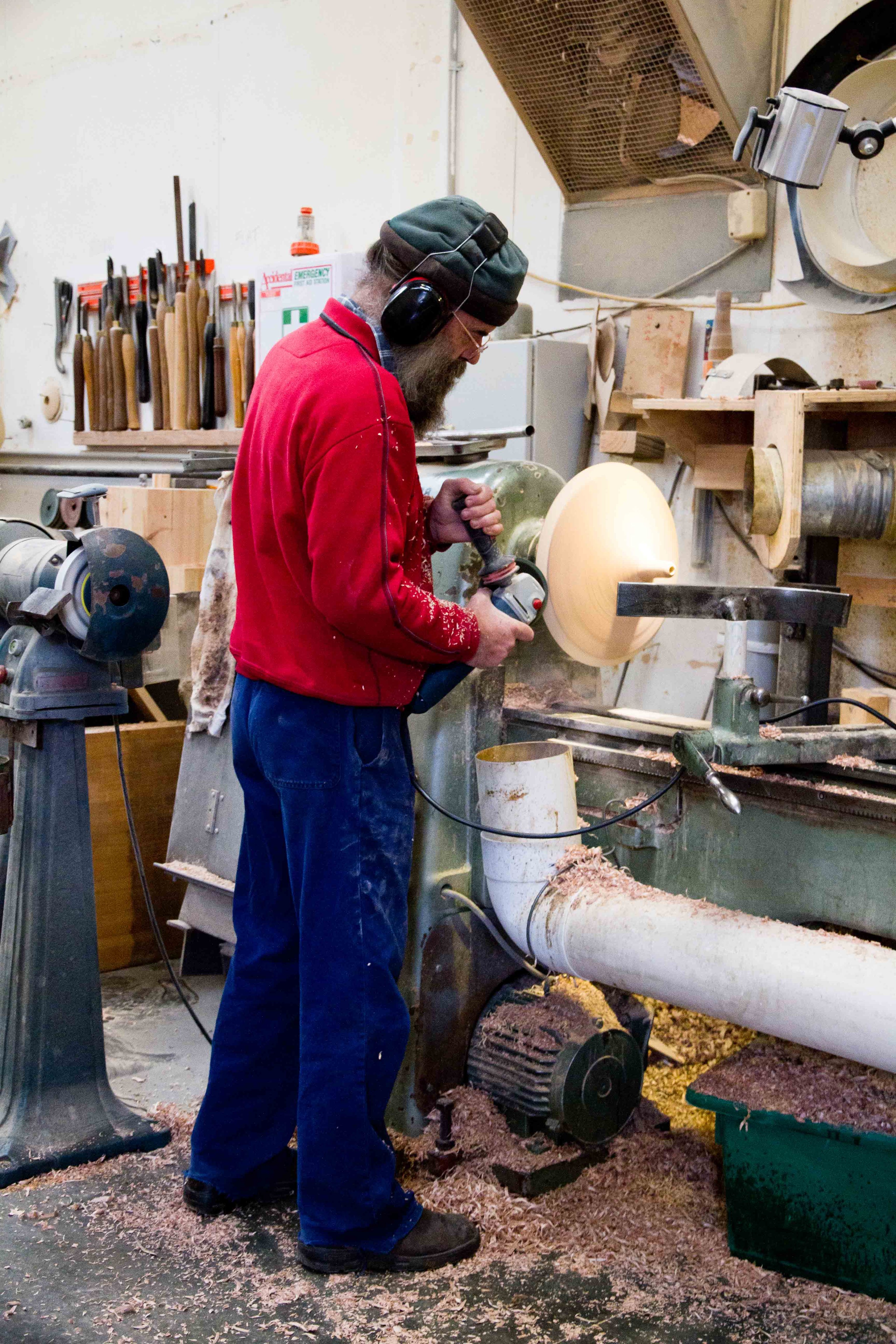 A man in a red jumper and blue pants standing in a timber workshop turning a Coco Pendant Mini timber top on a lathe. There are woodchips on the floor and timber turning tools hanging on the wall in background.